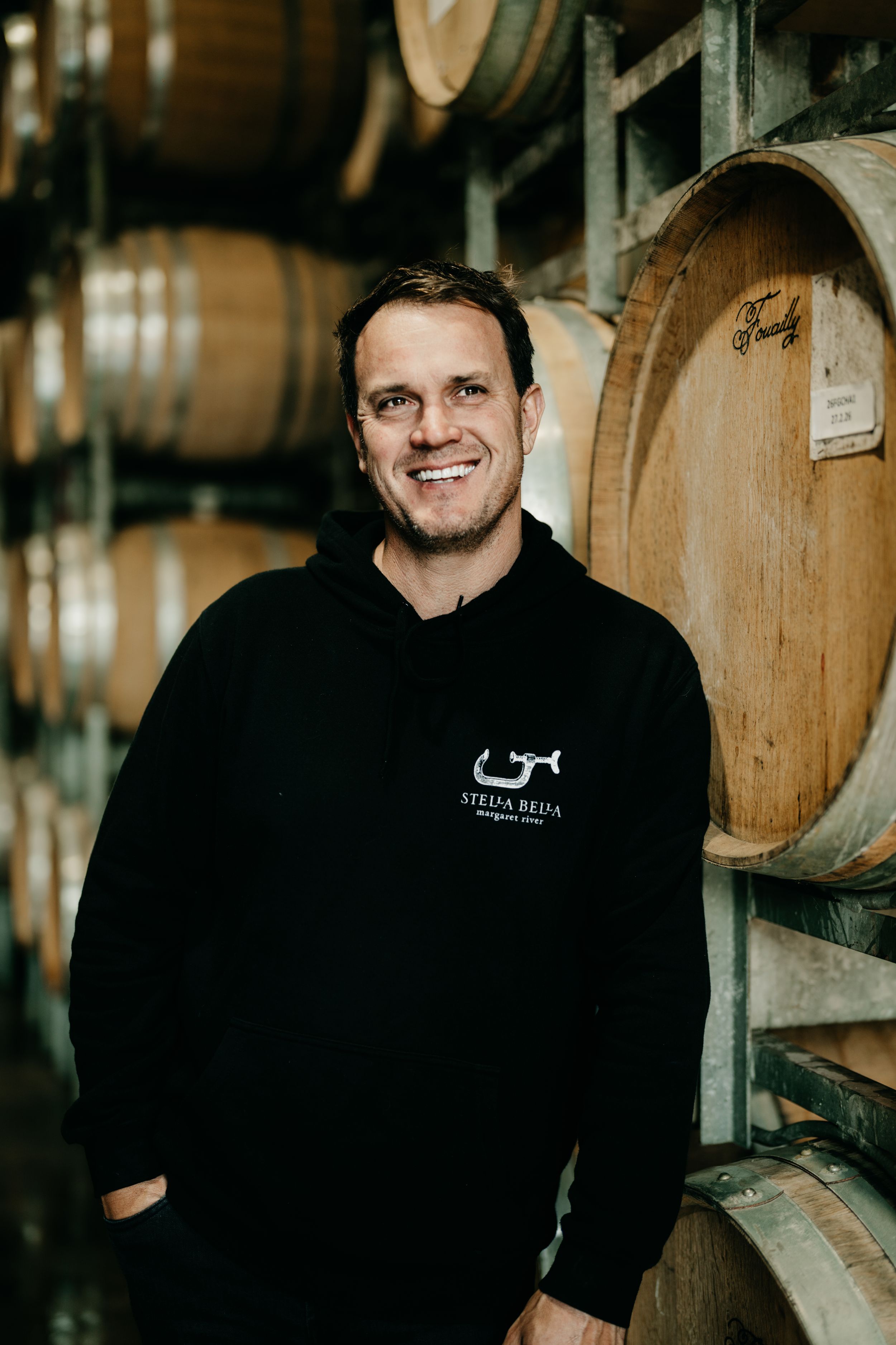 Smiling man in Stella Bella hoodie leaning against wine barrels in cellar.