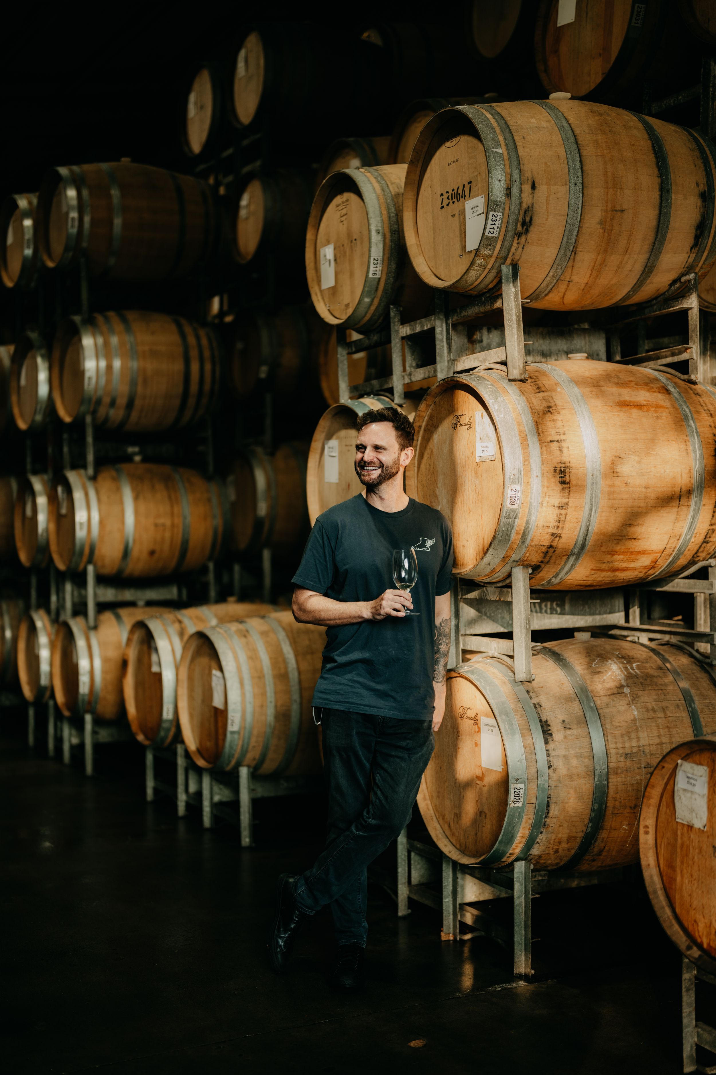 Man holding wine glass smiling among stacked oak barrels in a winery cellar.
