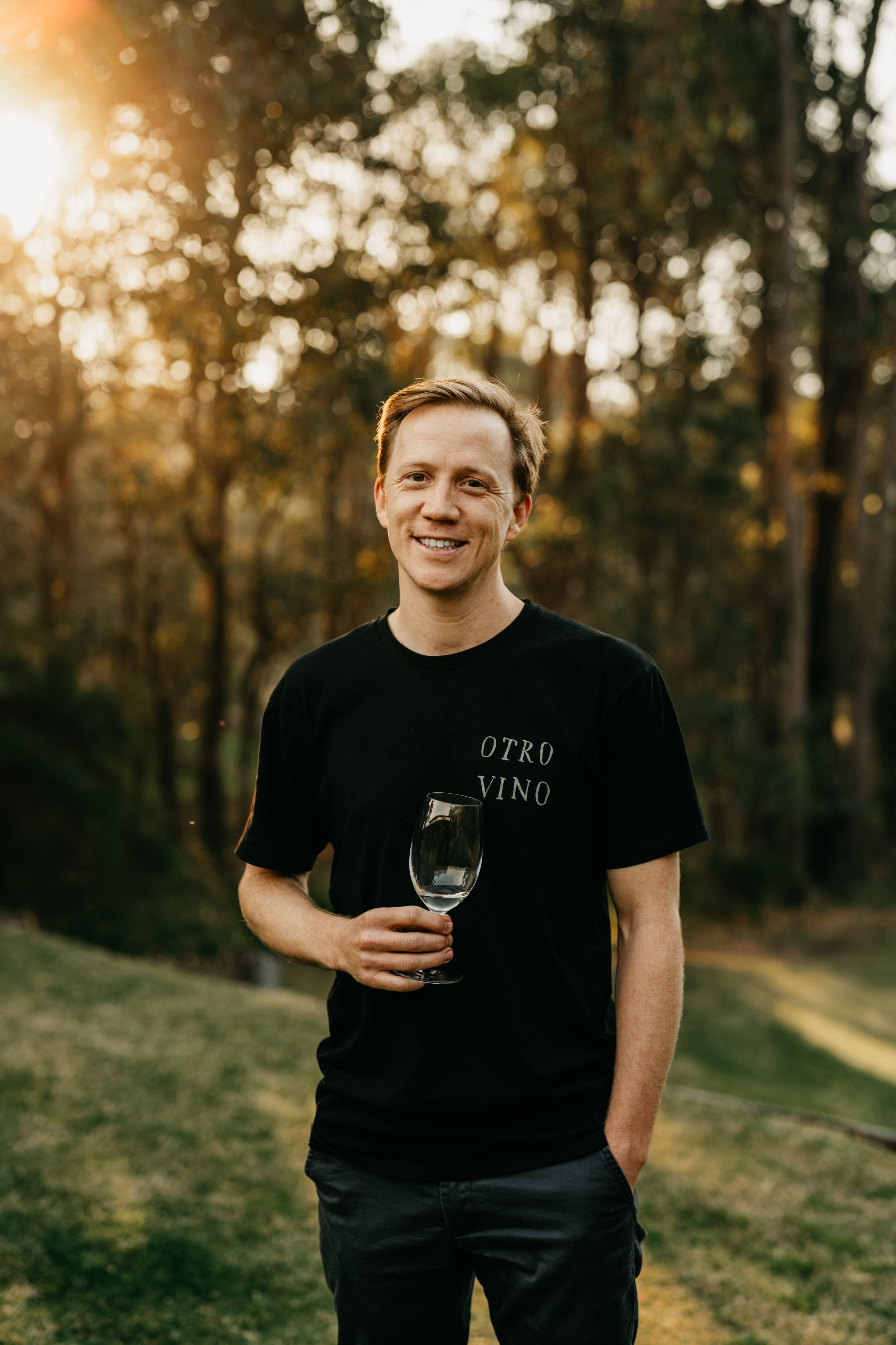 Smiling man holding a wine glass outdoors in golden sunlight wearing Otro Vino tee.