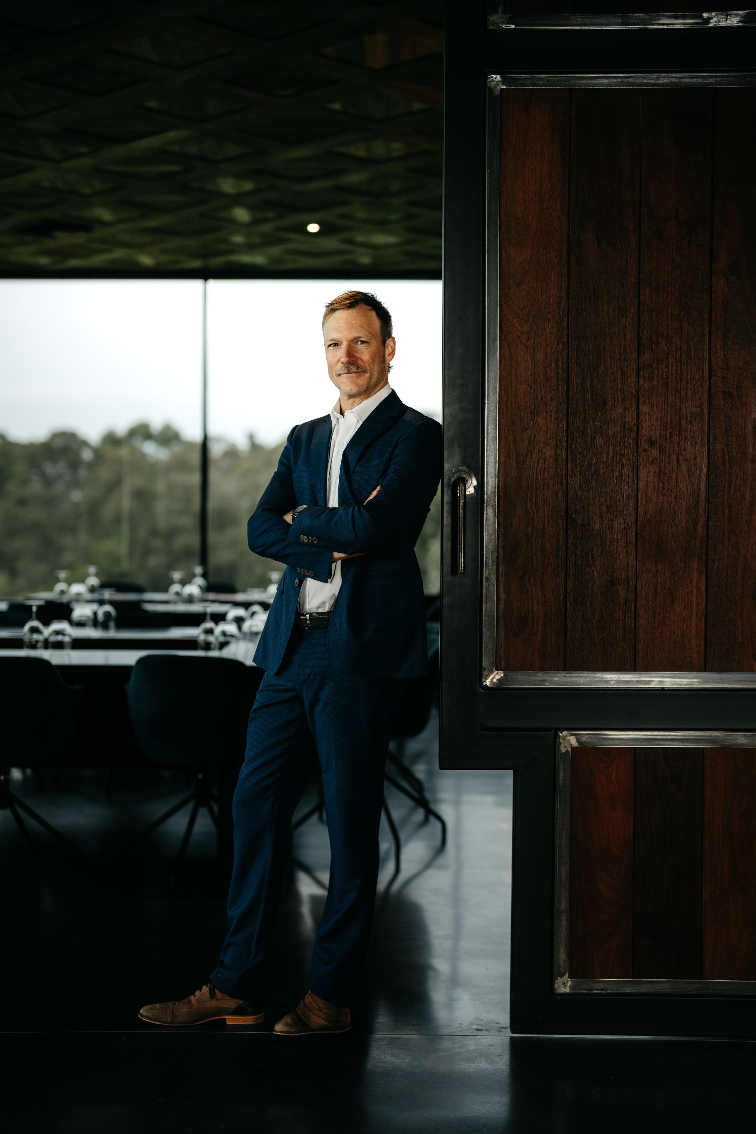 Confident businessman in navy suit standing with arms crossed beside a wooden door in a modern restaurant.
