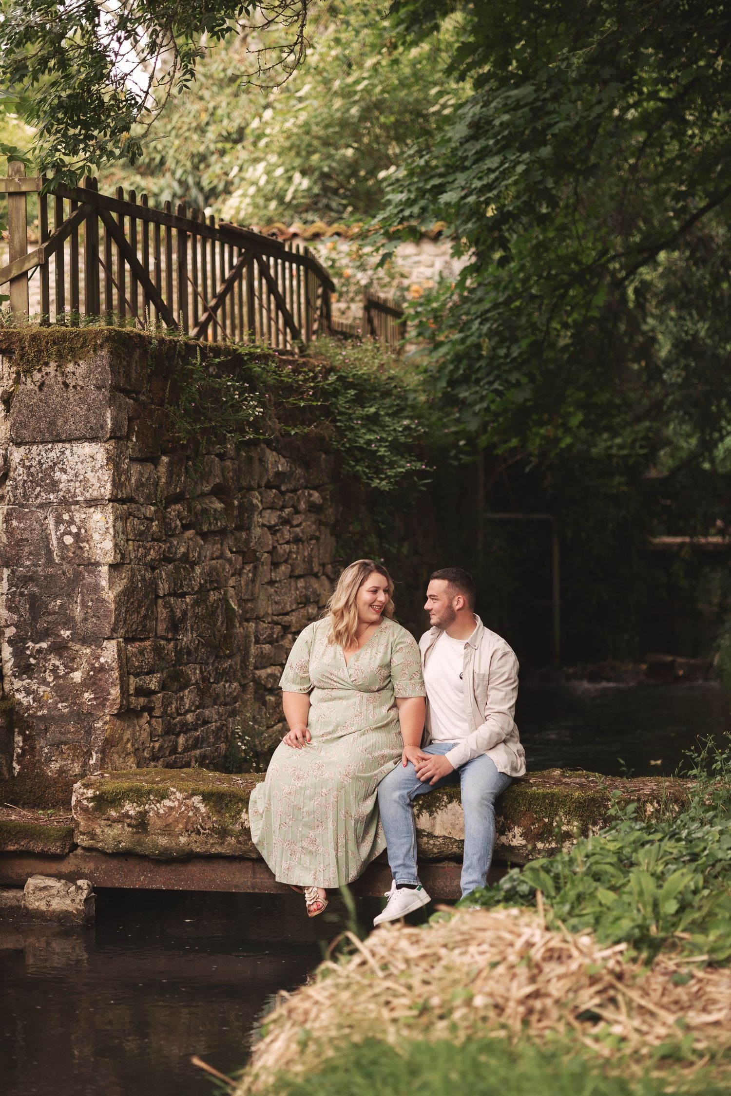 A couple in coordinating outfits with jeans and cardigans pose playfully back-to-back on a tree-lined nature path.