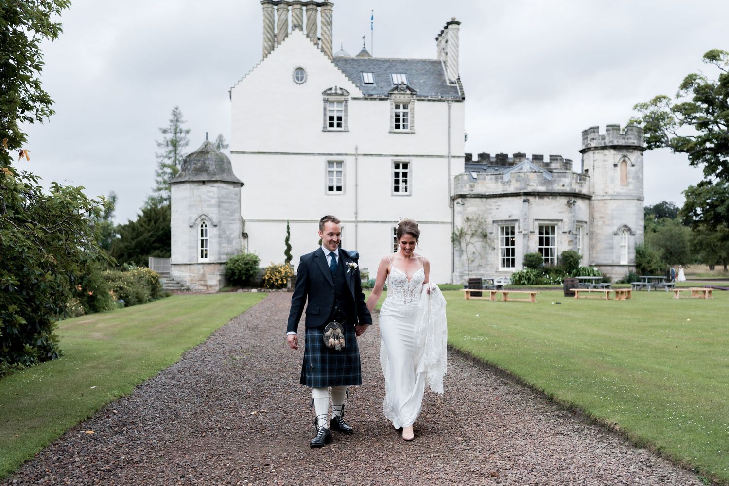 A couple walks together on a gravel path in front of a majestic white Scottish castle during their wedding celebration.