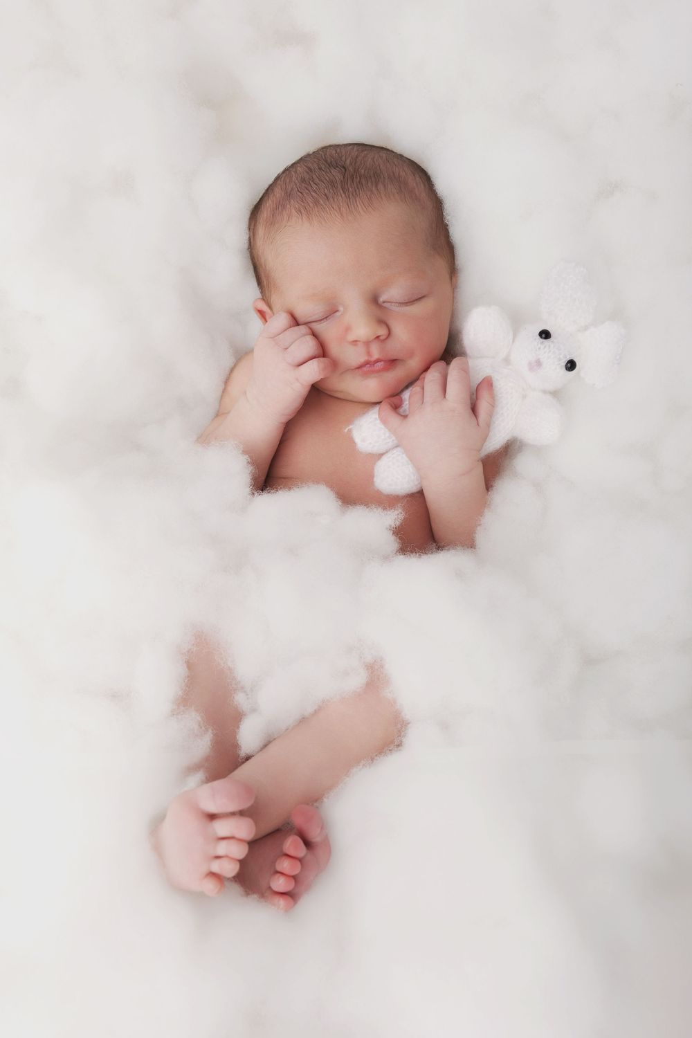 Peaceful newborn baby sleeping on white fluffy clouds, cuddling a tiny white stuffed bear toy.