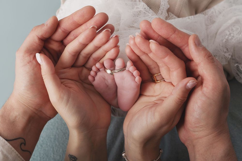 Two parents cradle their newborn baby's tiny feet in their hands, both wearing wedding rings, on a soft gray background.
