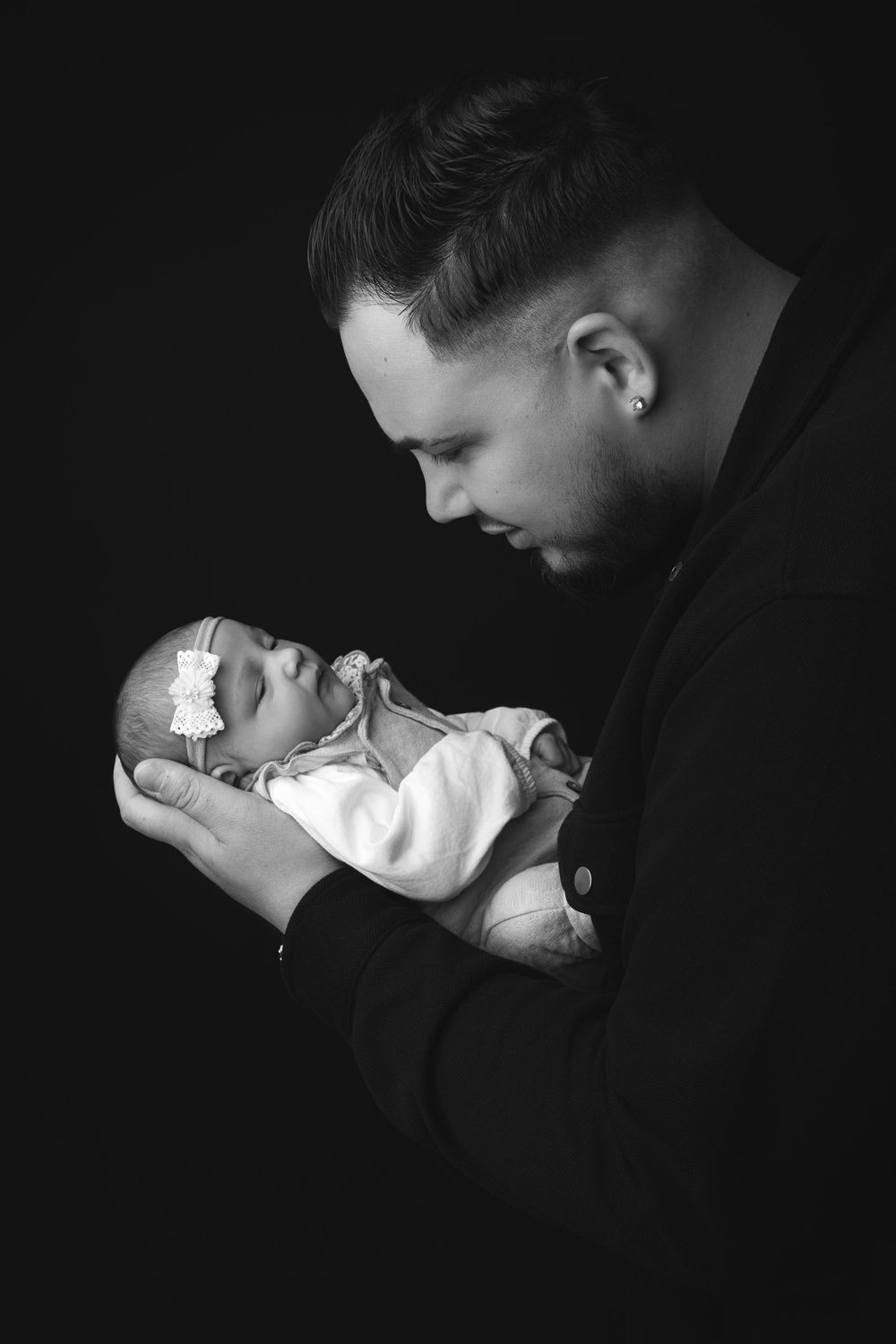 Black and white portrait of a father tenderly holding his sleeping newborn baby girl against a dark background.