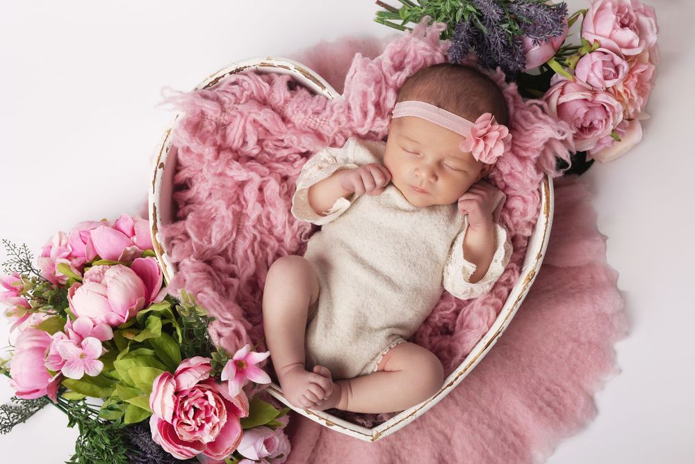 Newborn baby sleeping peacefully in a heart-shaped basket with pink flowers and a floral headband.
