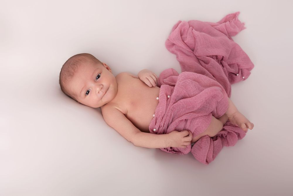 Newborn baby wrapped in a soft pink muslin cloth, lying on a white background, smiling peacefully.