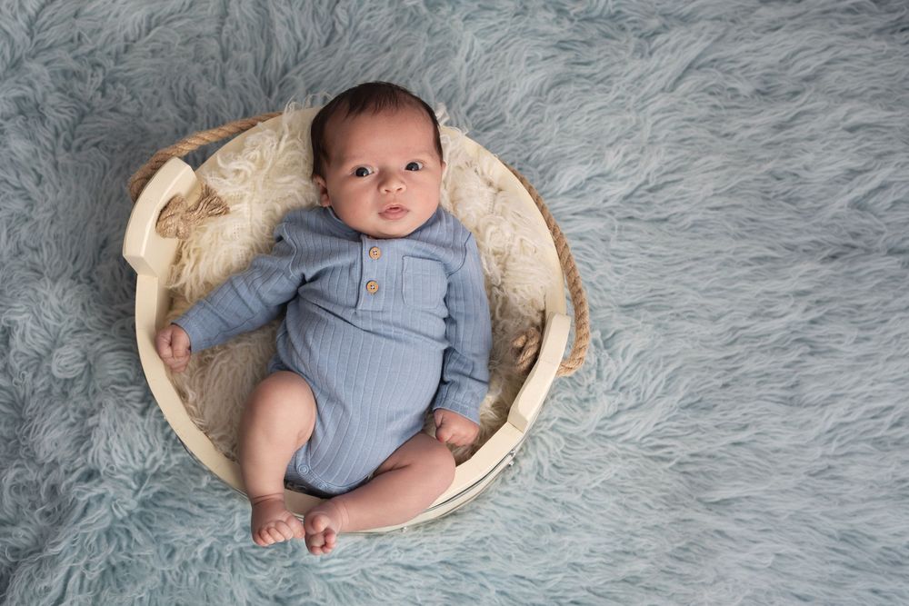 Newborn baby in blue romper lying in wooden bowl on soft gray fur blanket during professional photo session.