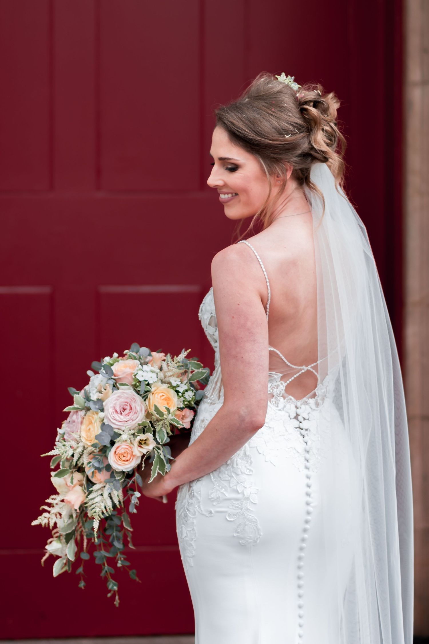 A bride in a fitted white lace gown with button details holds a peach and blush rose bouquet against a red door.