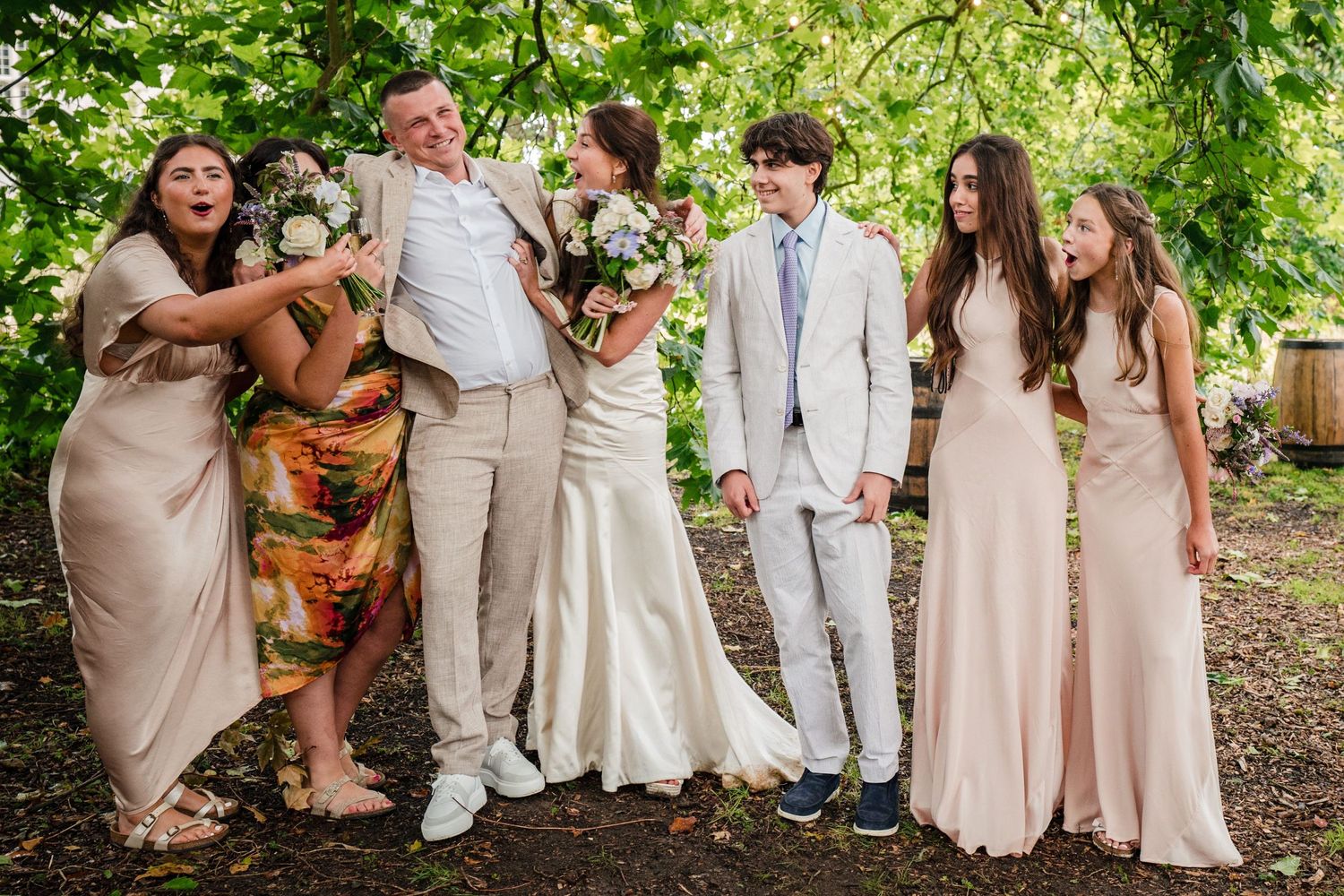 A joyful wedding party poses together outdoors under green trees, wearing coordinating blush and neutral-toned attire in Nova Scotia photo.