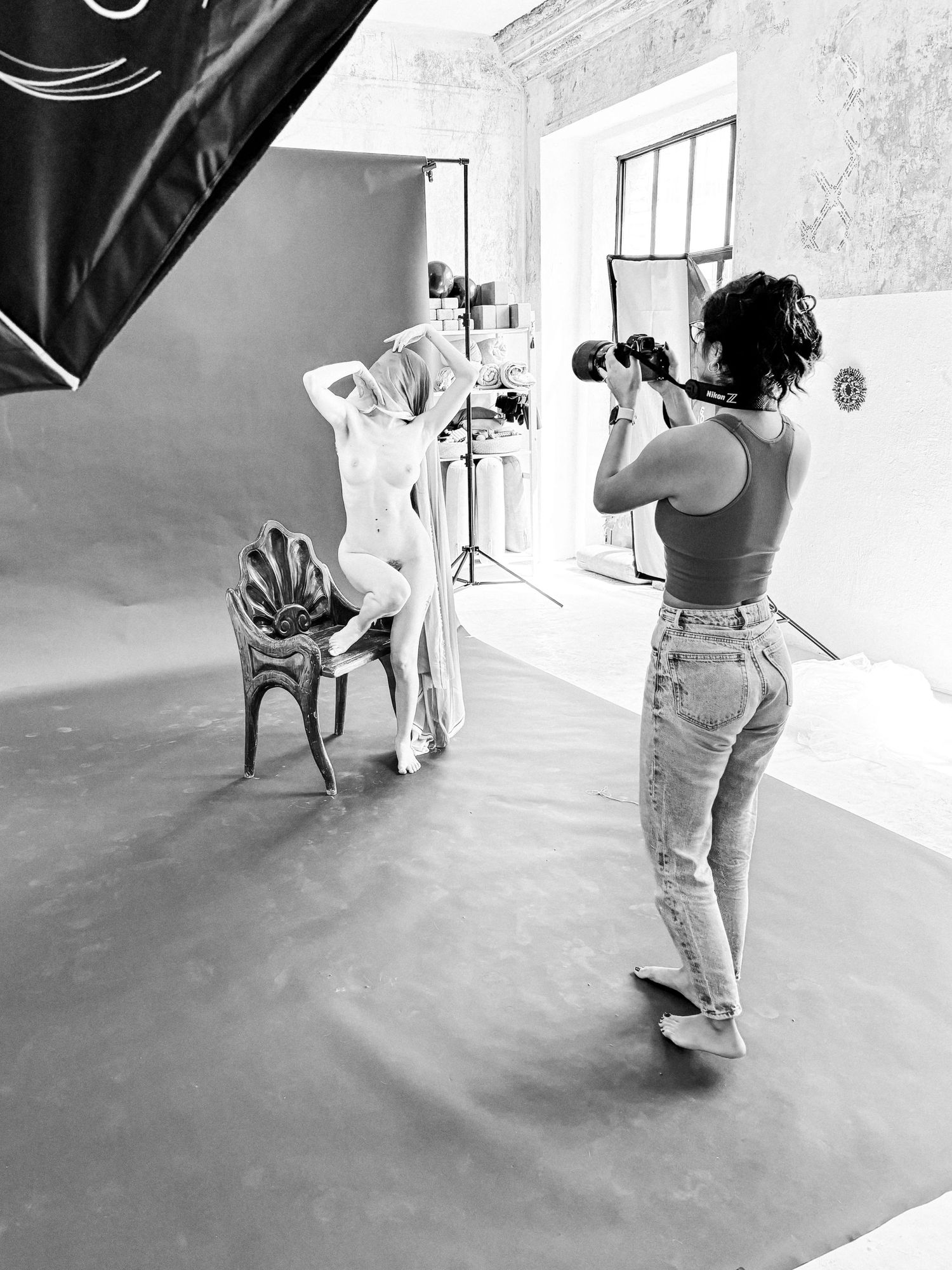 A black and white photo of a photography session in a studio with a model posing on a chair and photographer at work.