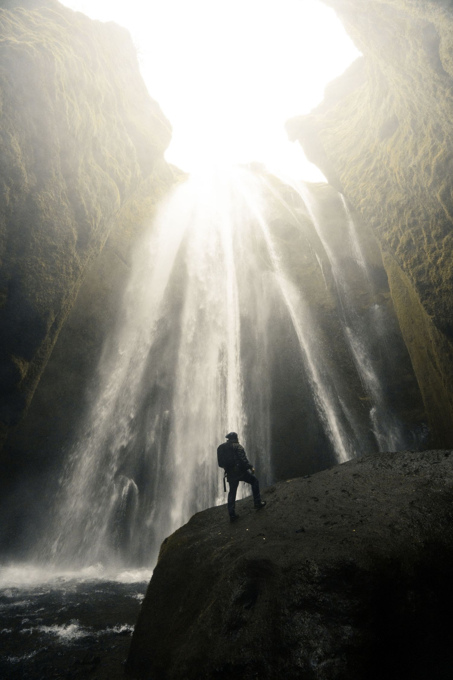 Silhouette of hiker beneath majestic waterfall with sunlight streaming through.