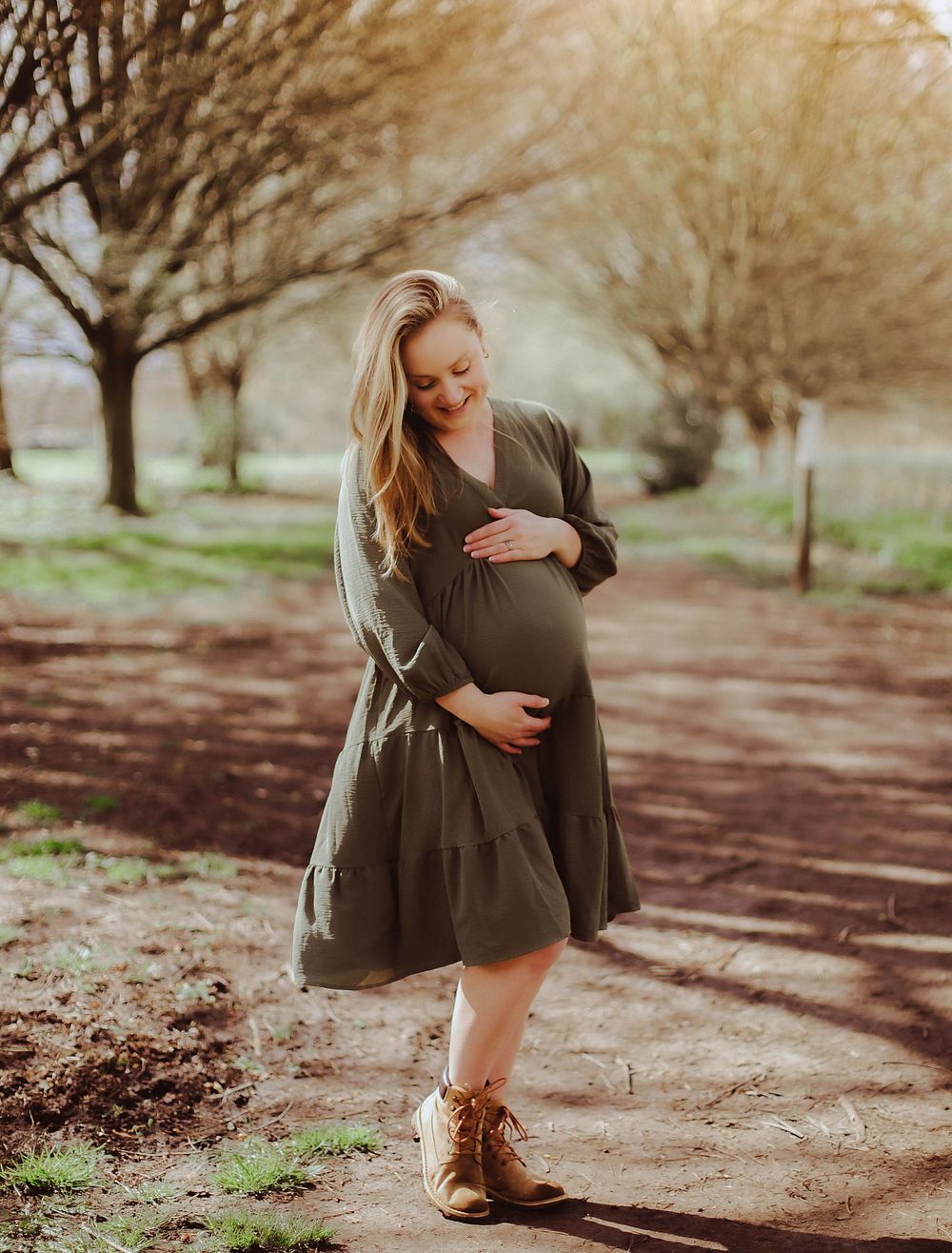 A pregnant woman in a brown dress and boots poses along a tree-lined path on an autumn day.
