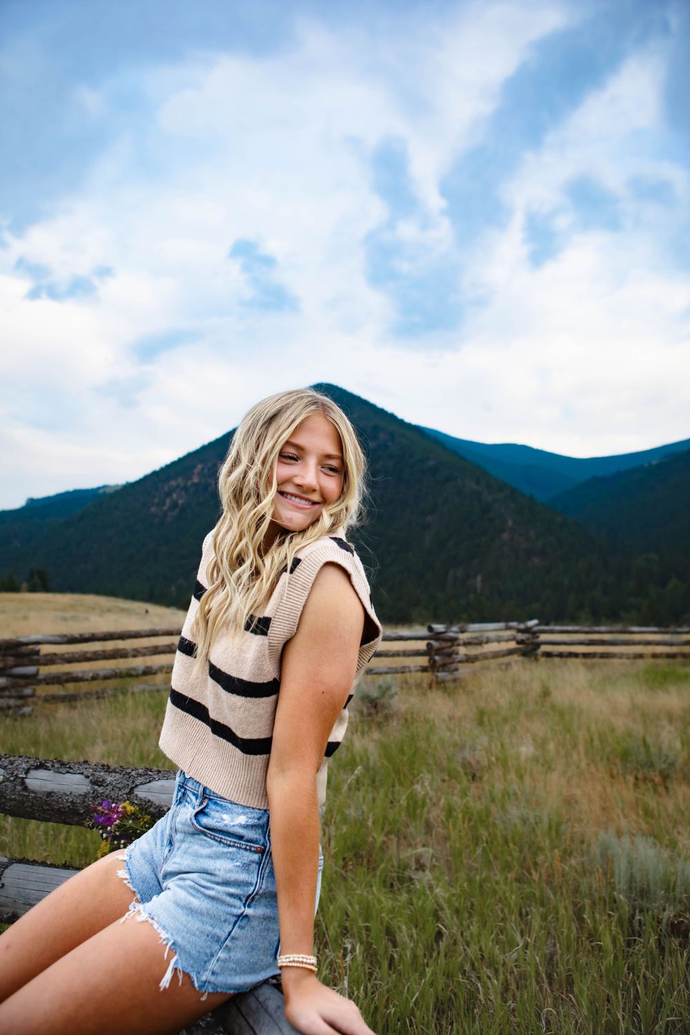 A person in a striped tank top and denim shorts sits on a rustic fence with mountains and blue sky in the background.