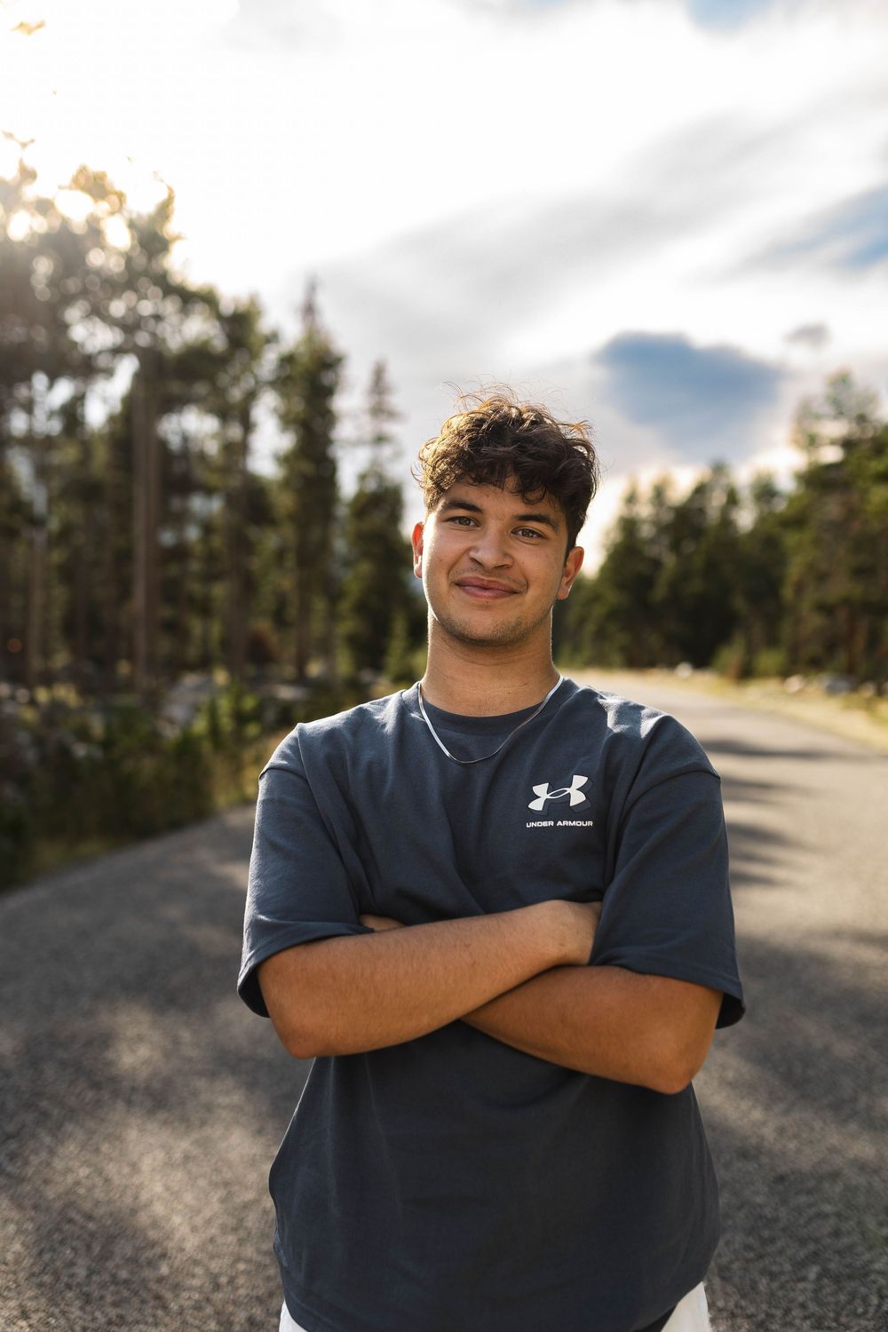 A person in a navy Under Armour shirt smiles while standing on a gravel road with trees and sky in the background.