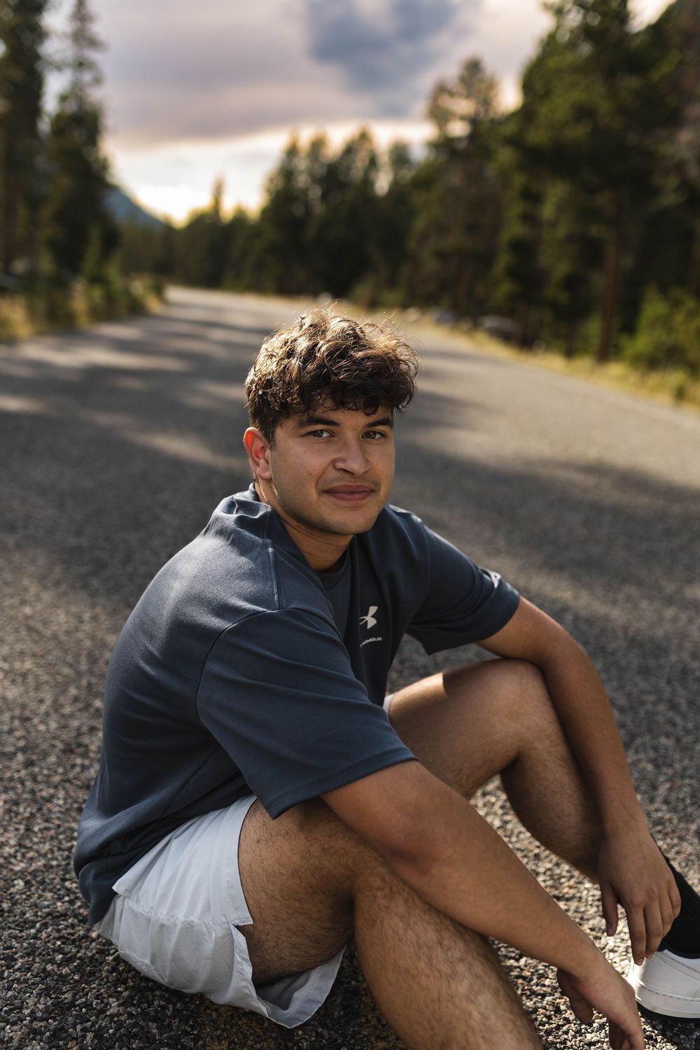 Person sitting on asphalt road surrounded by evergreen trees during golden hour.