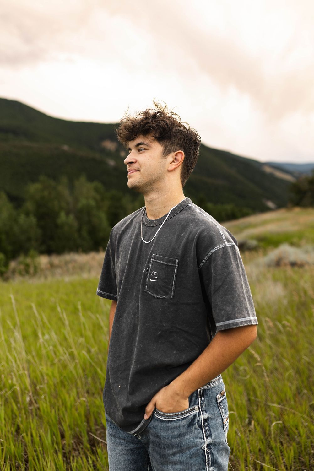 Person standing in tall grass meadow with mountains and sunset sky in background.