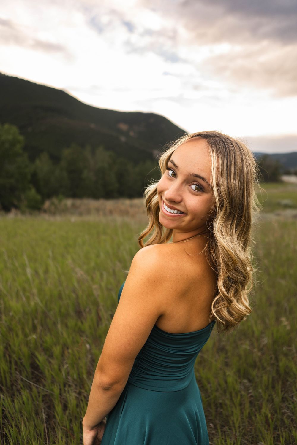 A person in a teal dress poses in a field at sunset with mountains in the background.