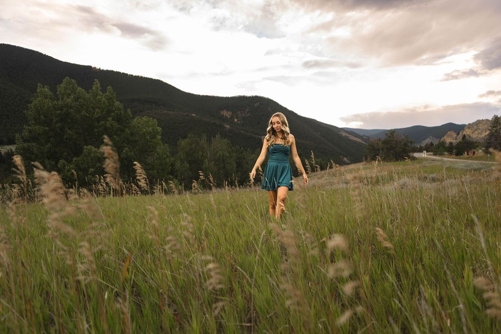 A person in a flowing dress walks through a golden meadow at sunset with mountains in the background.