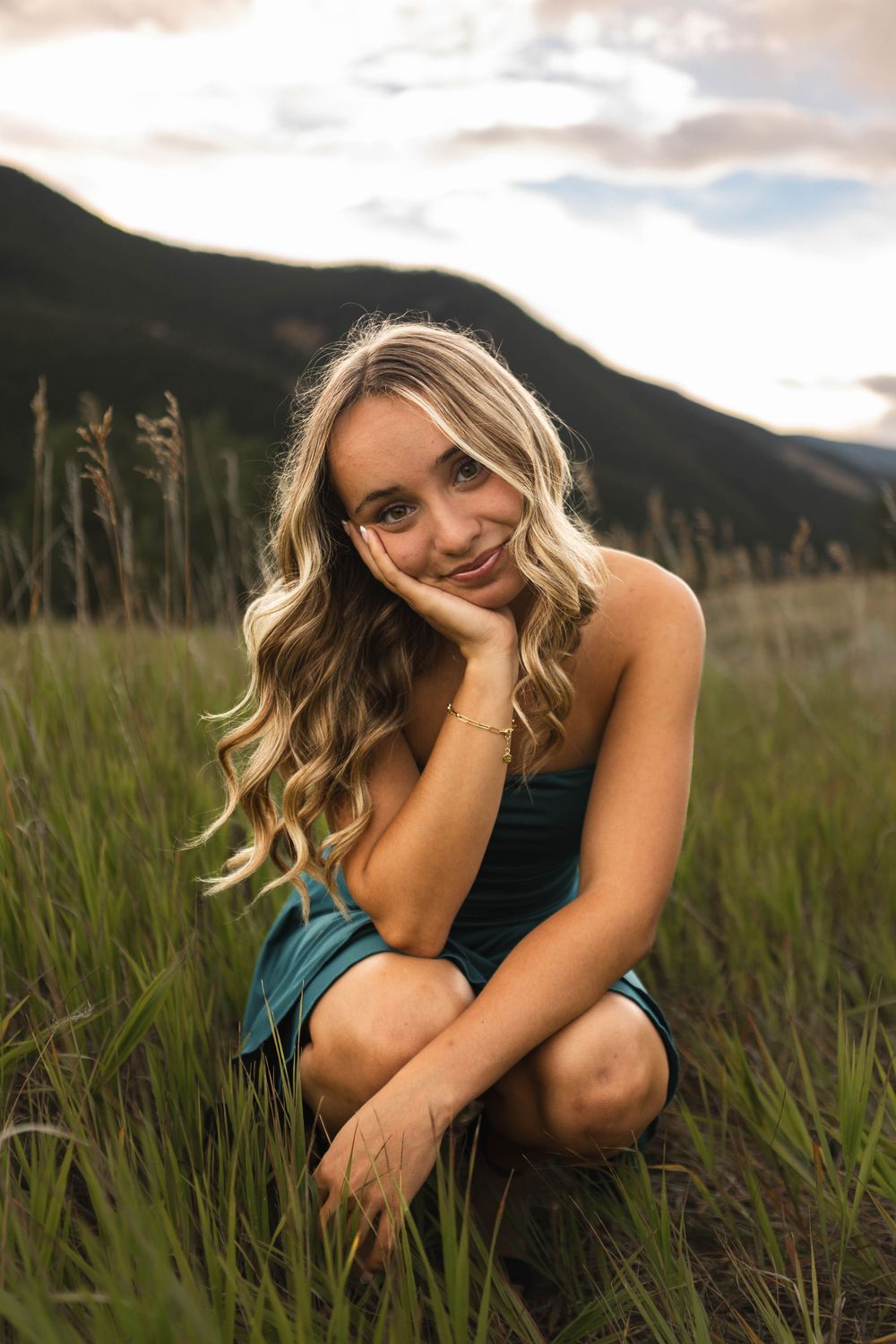 A person sits in tall grass at sunset in a contemplative pose with mountains visible in the background.