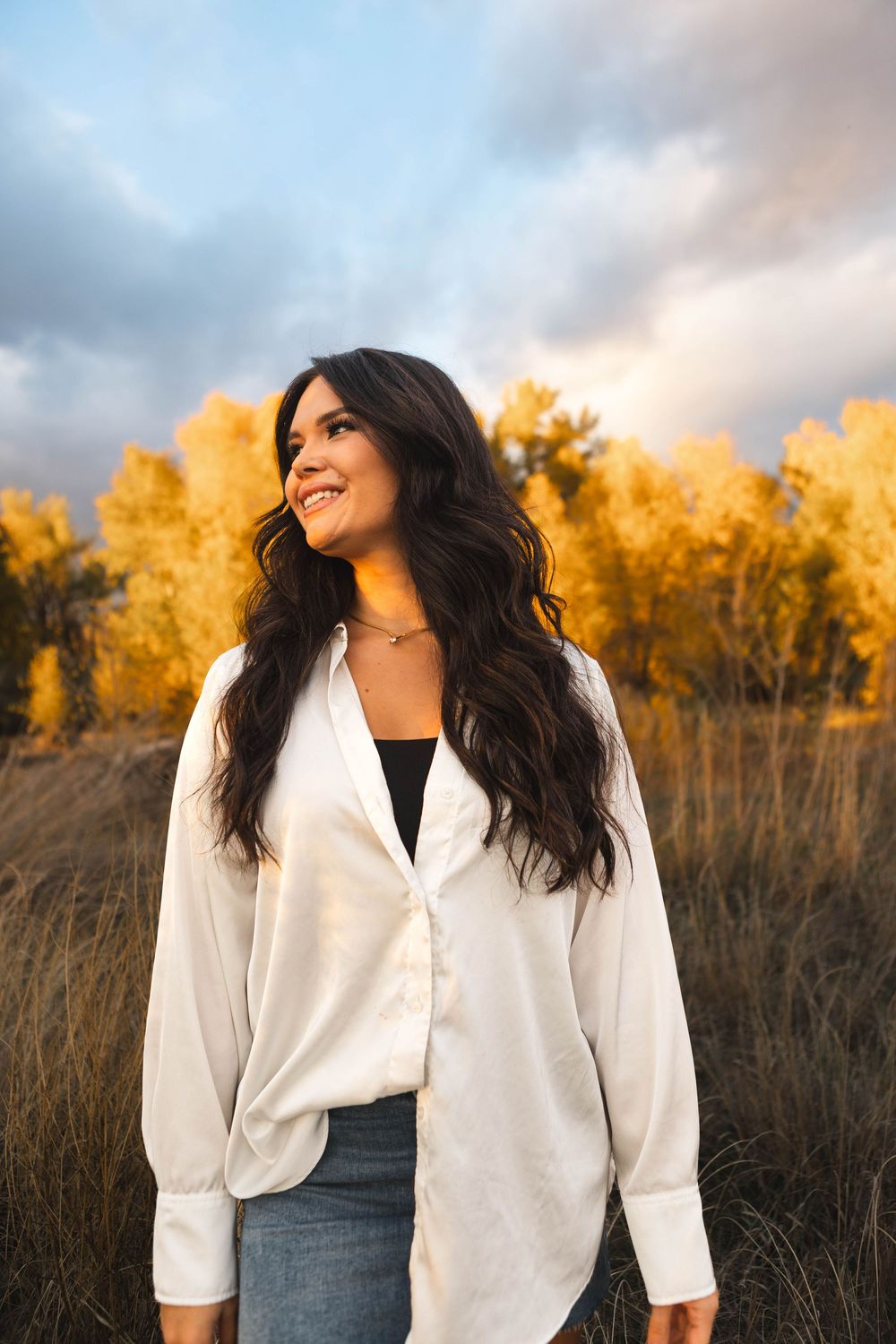 A woman in a white button-down shirt smiles while standing in a field with golden autumn trees in the background.