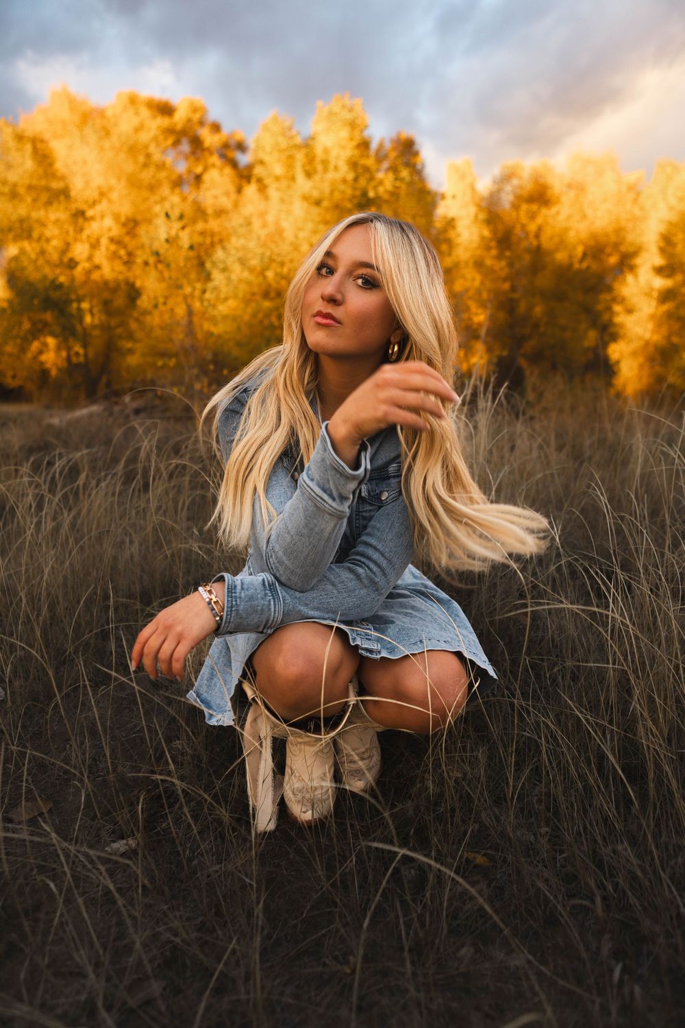 A figure in a denim jacket poses in tall grass at sunset with golden autumn trees in the background.