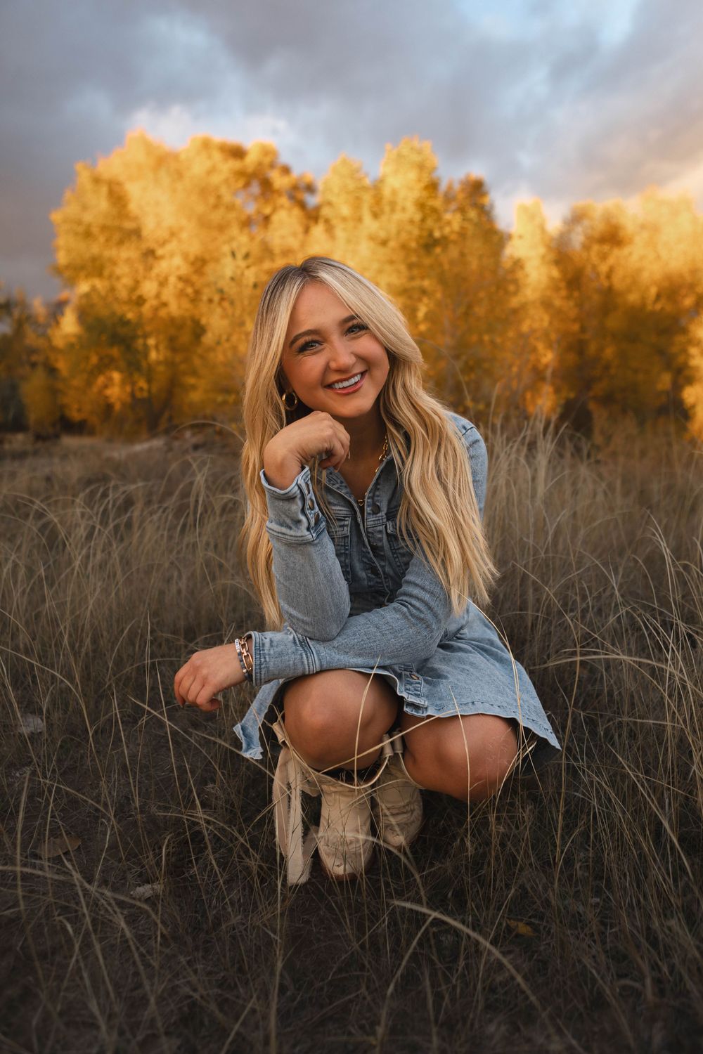 A person in a denim dress crouches in a field of tall grass during golden hour with autumn trees in the background.