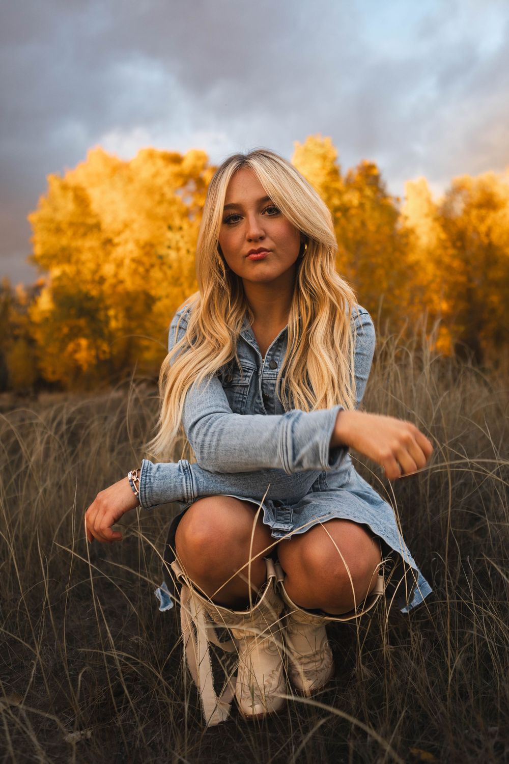 A person in denim crouches in tall grass during golden hour as autumn trees glow orange in the background.