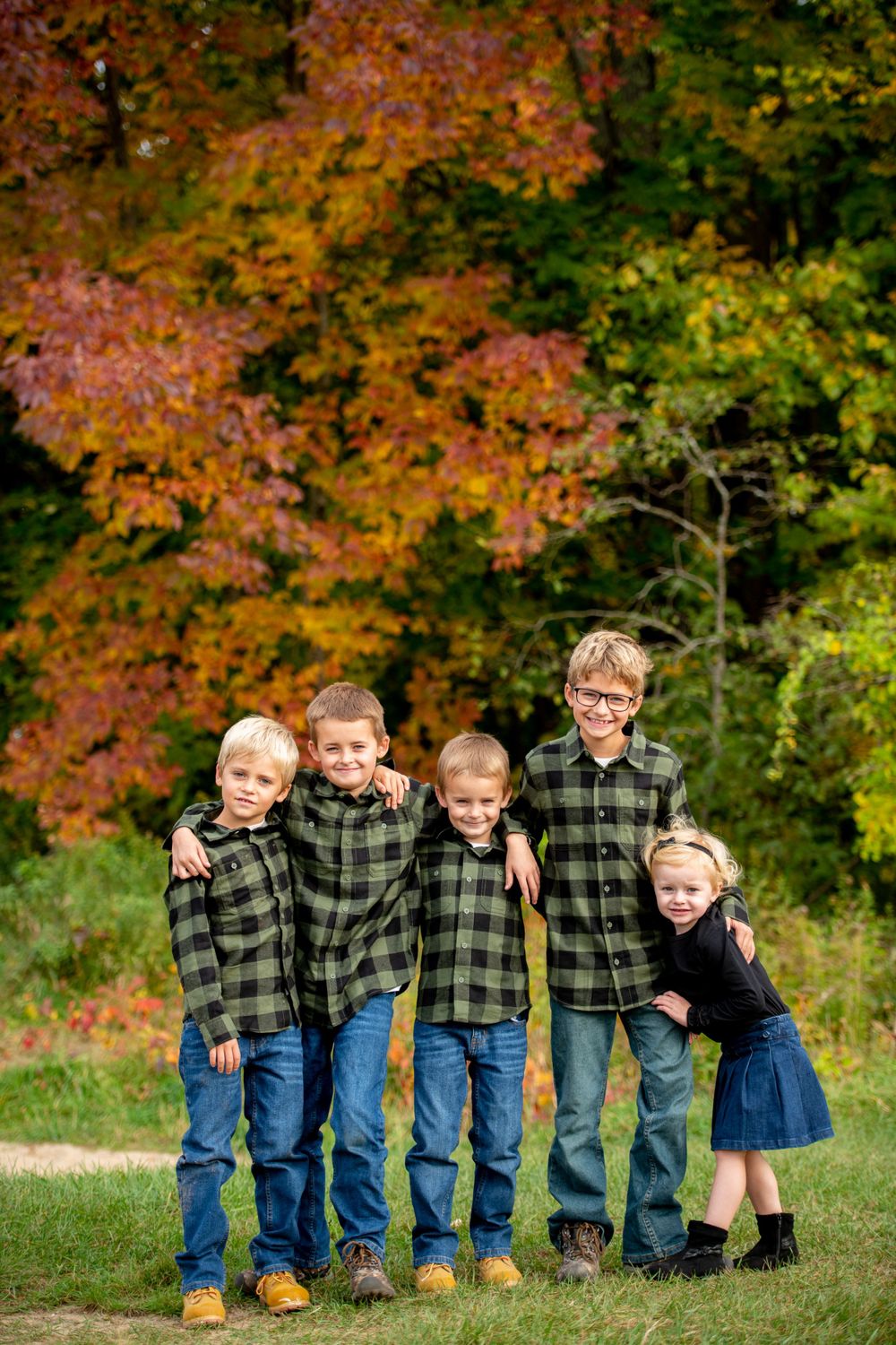 Children wearing matching plaid shirts and jeans pose together against vibrant autumn foliage.
