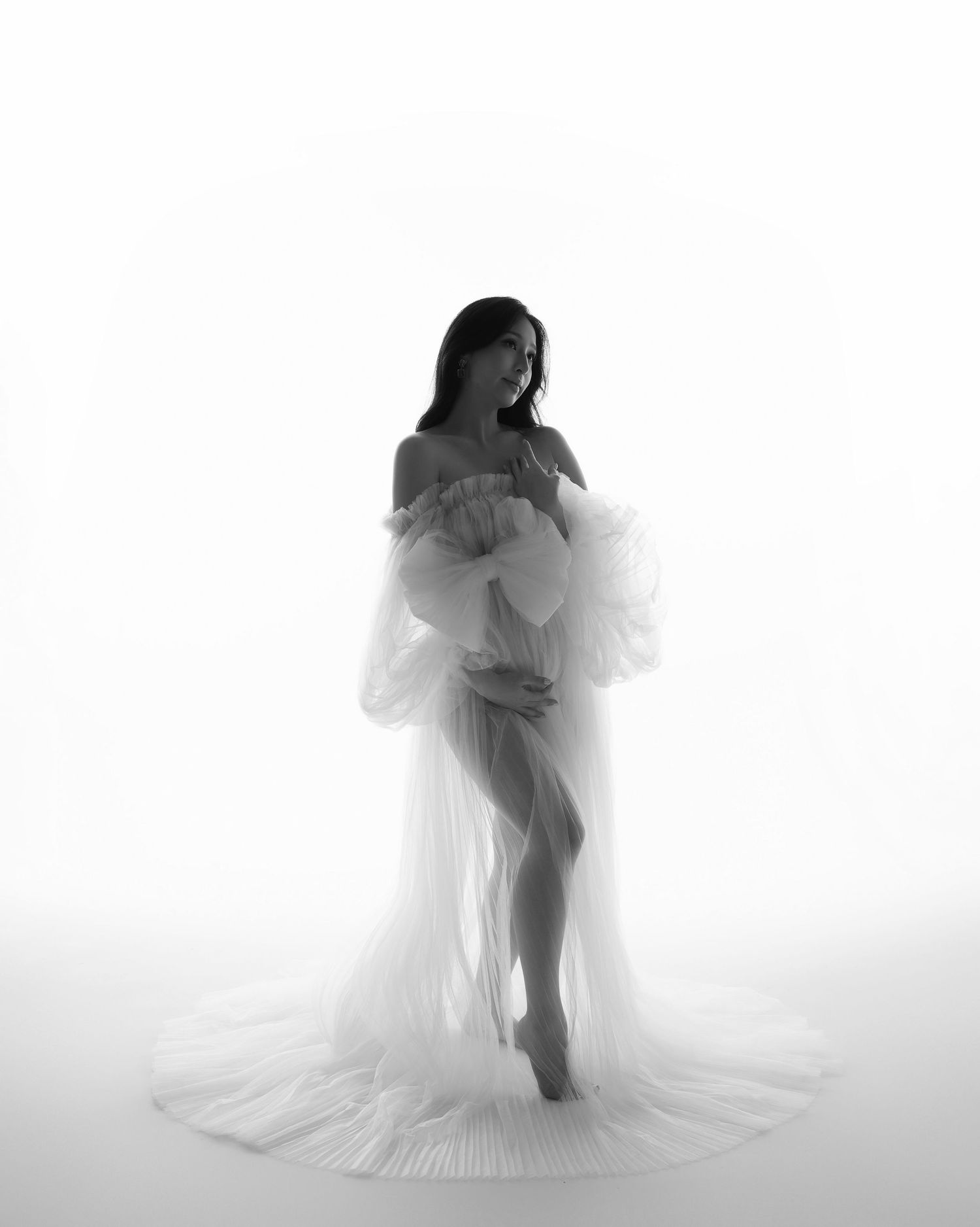 Artistic black and white maternity silhouette portrait with flowing sheer fabric against a bright white background.