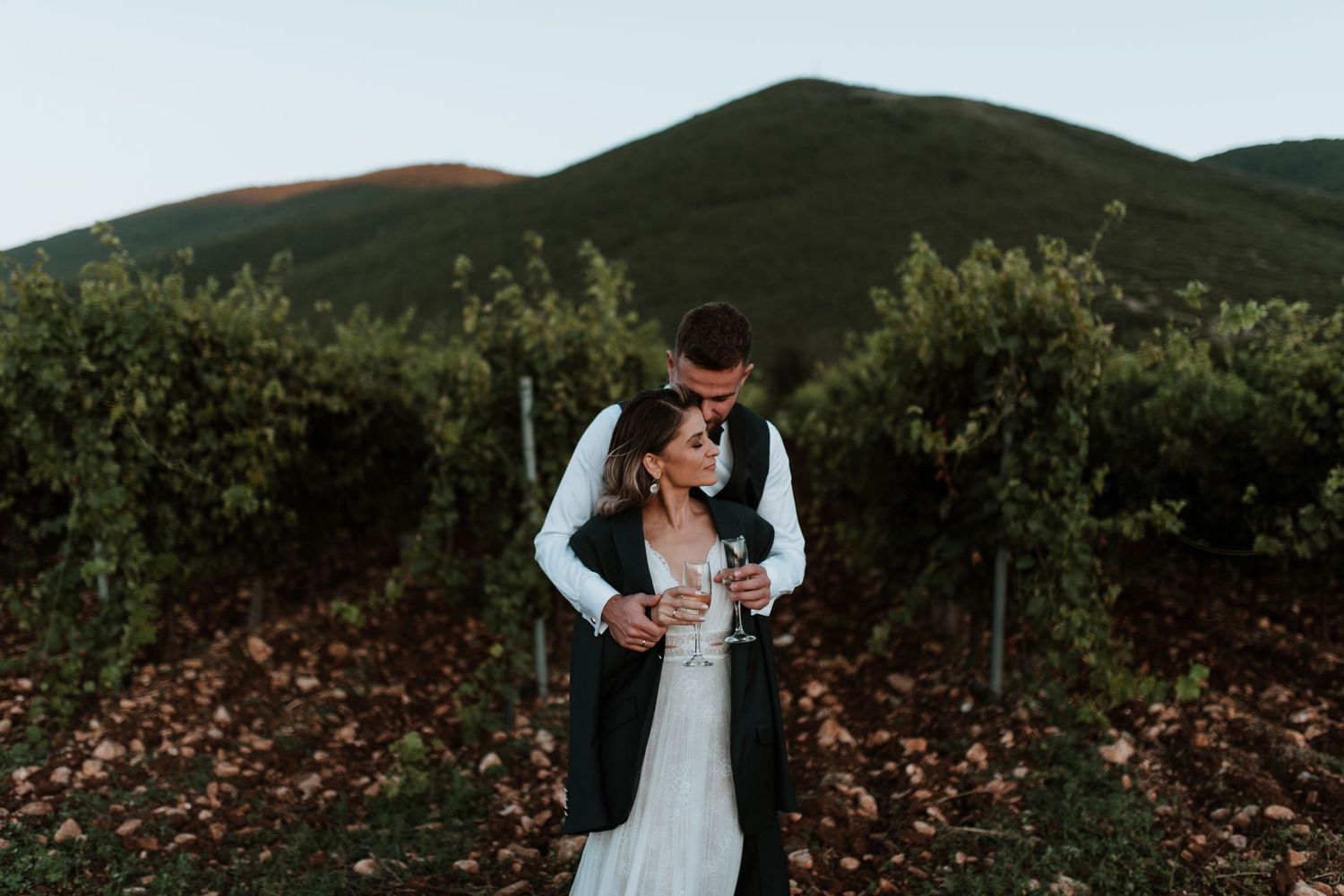 A couple shares a tender moment among vineyard rows with mountains behind them.