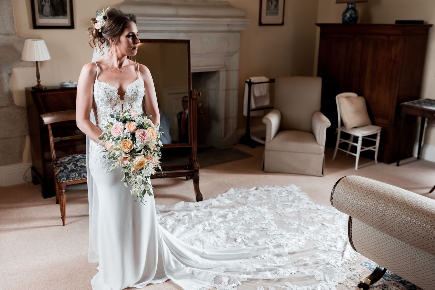 Wedding dress with long lace train and pastel bouquet in an elegant interior room with vintage furniture.
