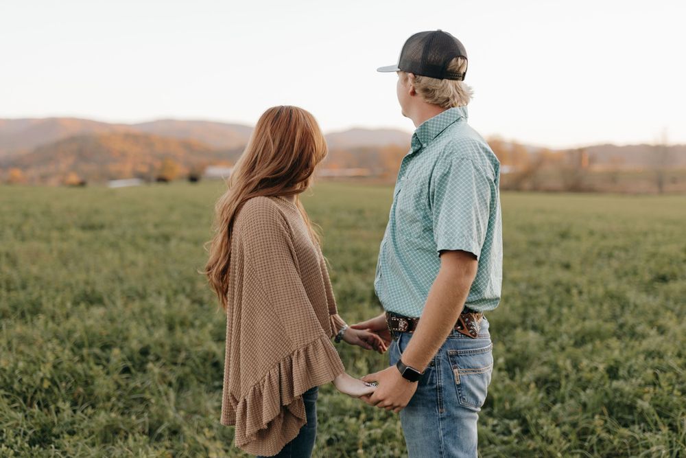 A couple holds hands while standing in a green field with rolling hills in the background.