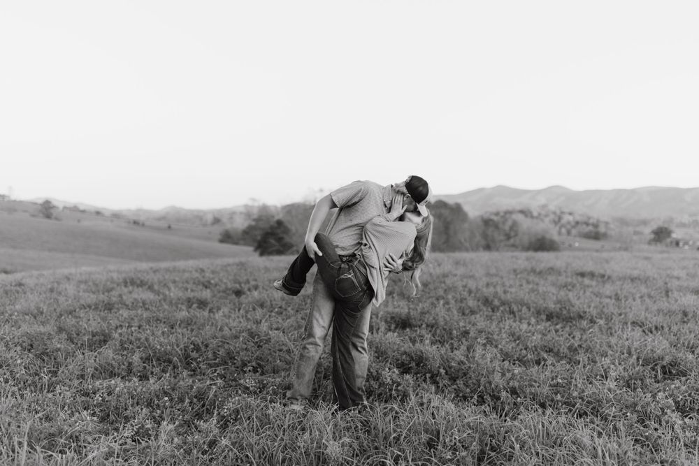 A couple embraces in a dramatic lift pose against a scenic hillside landscape.