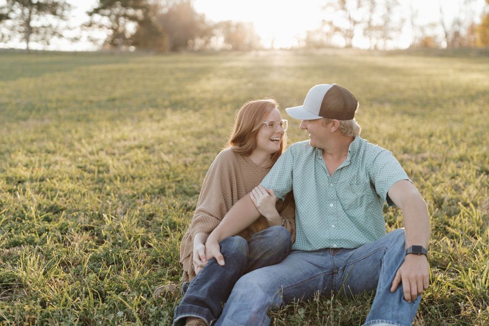 A couple sits together on grass during sunset, sharing a laughing moment.