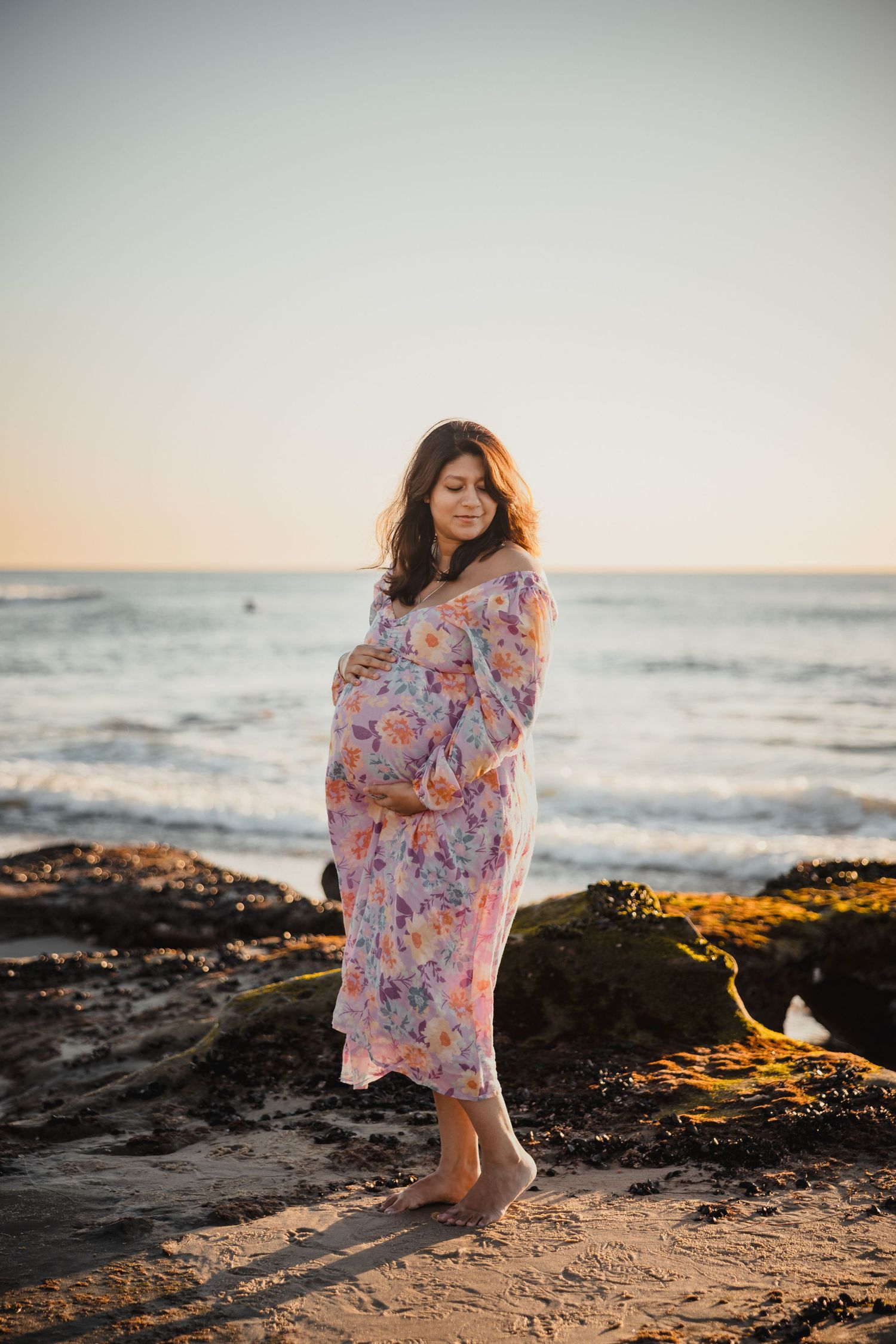 Pregnant woman in floral dress stands barefoot on beach at sunset with ocean waves in background.