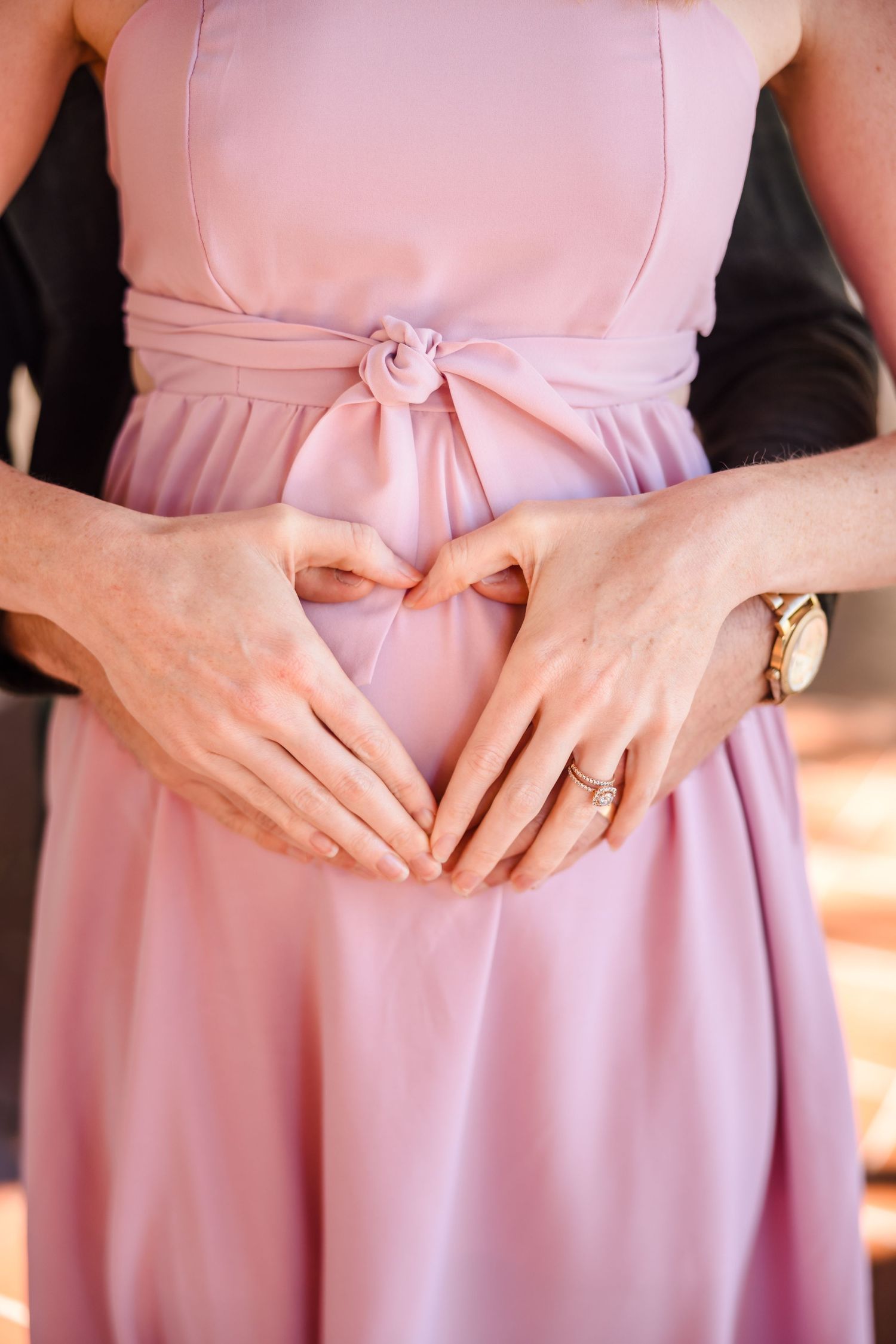 Hands form a heart shape over a baby bump while wearing a pink maternity dress with a bow sash.