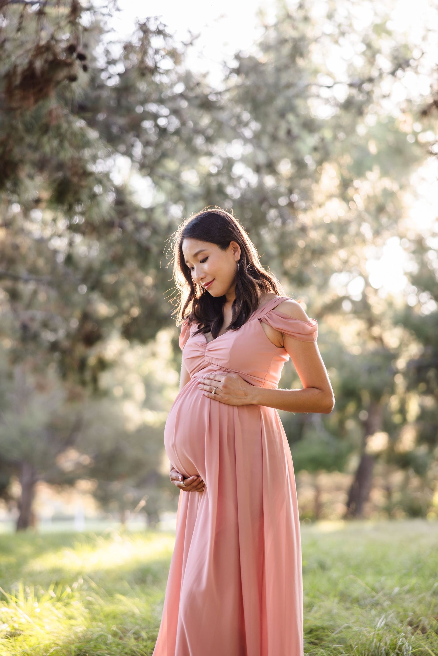 Woman in a flowing pink maternity dress stands in a sunlit outdoor setting with trees in the background.