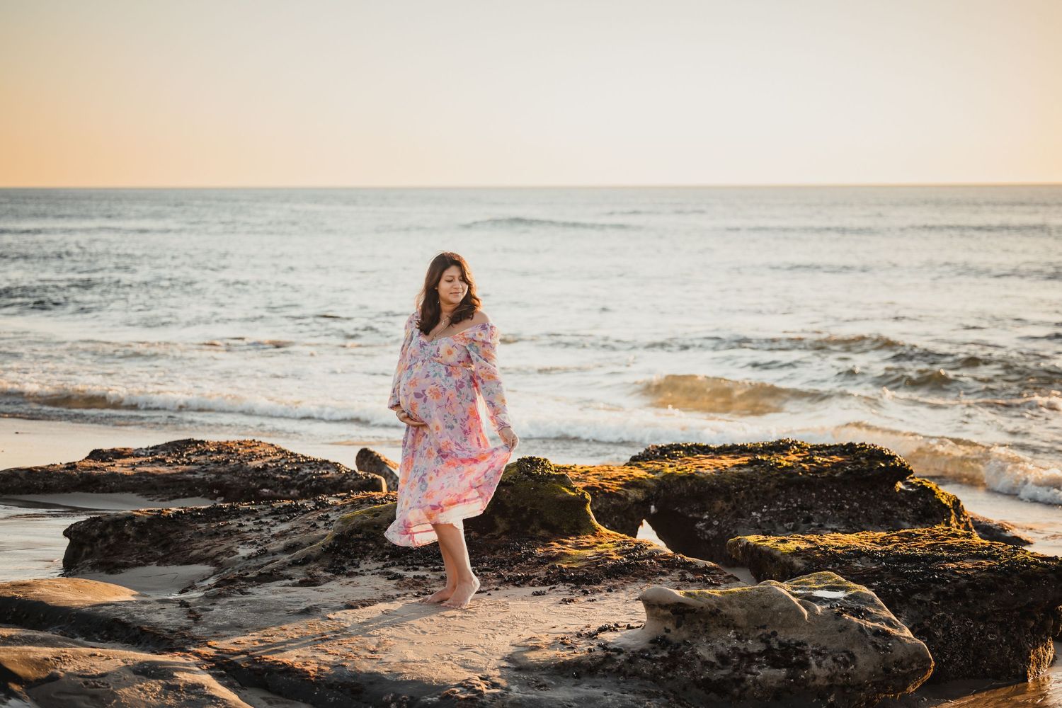 Someone in a pink floral dress sits on coastal rocks at sunset as ocean waves crash against the shore.