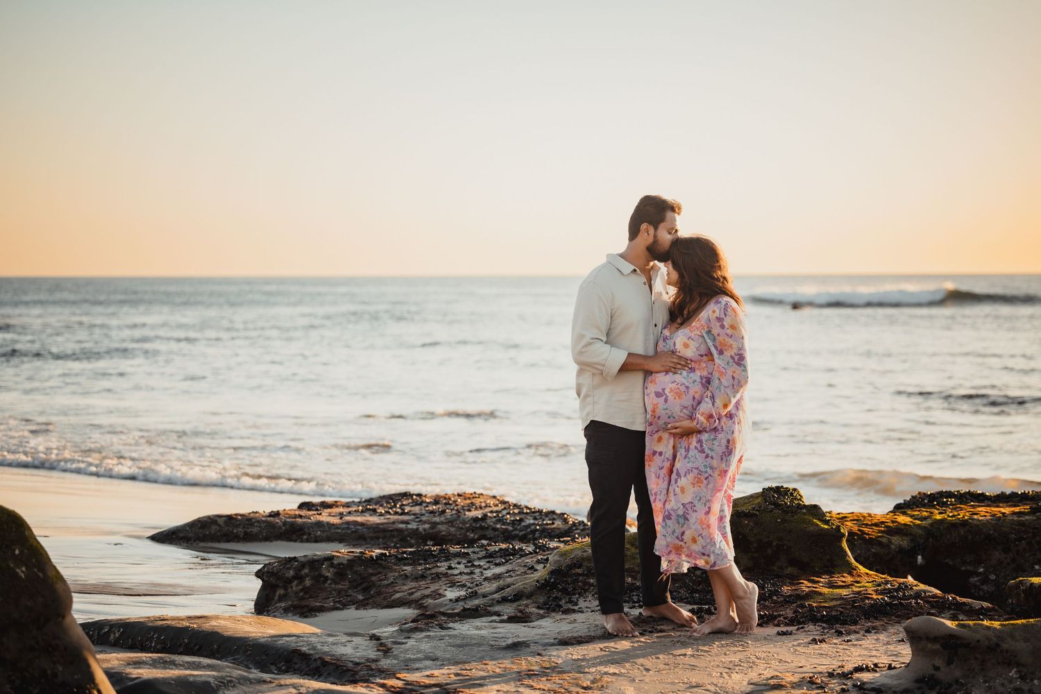 A couple stands together on rocky beach during golden hour sunset as waves crash against the shoreline.