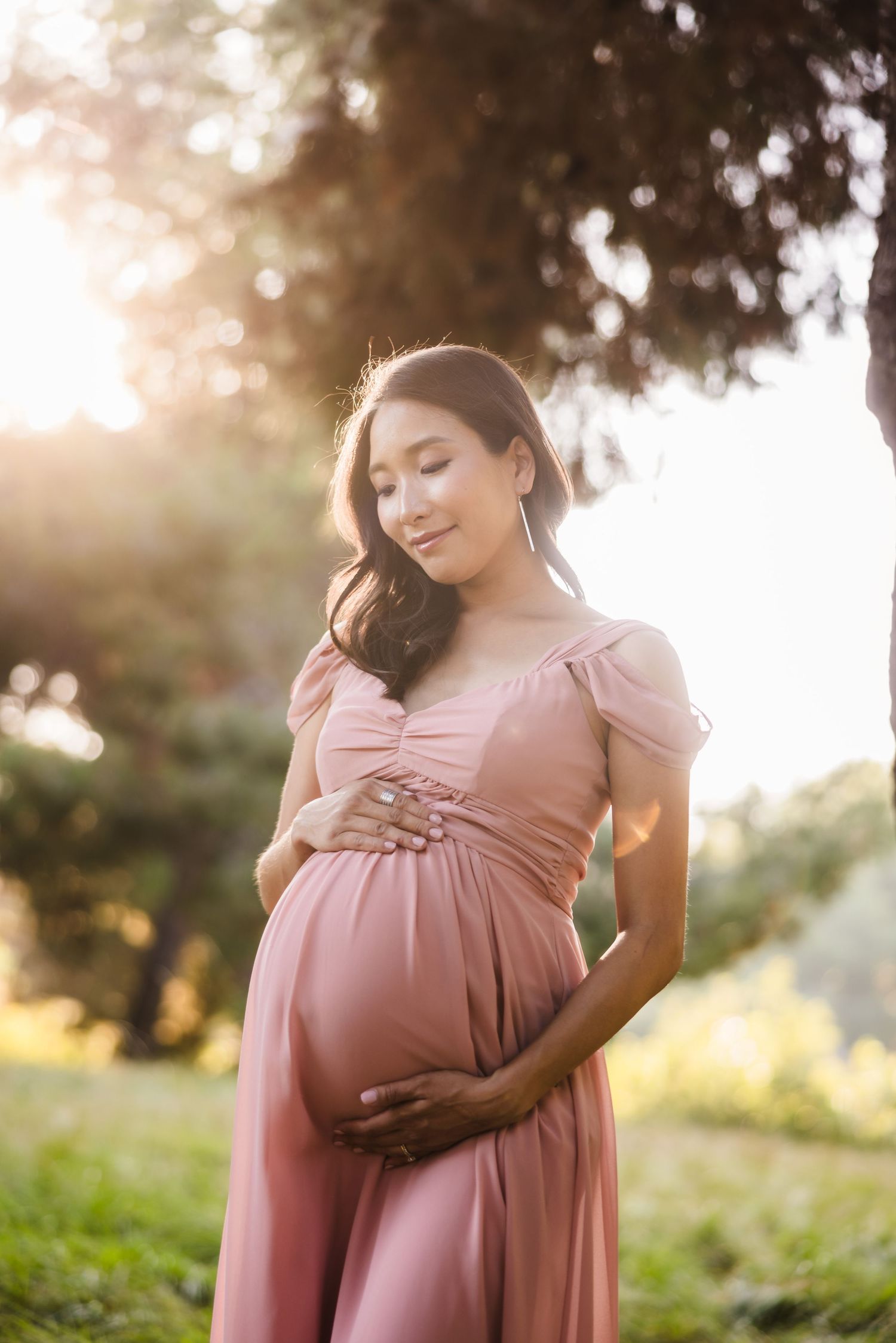 A pregnant woman in a pink dress poses outdoors during golden hour sunset with her hands on her belly.