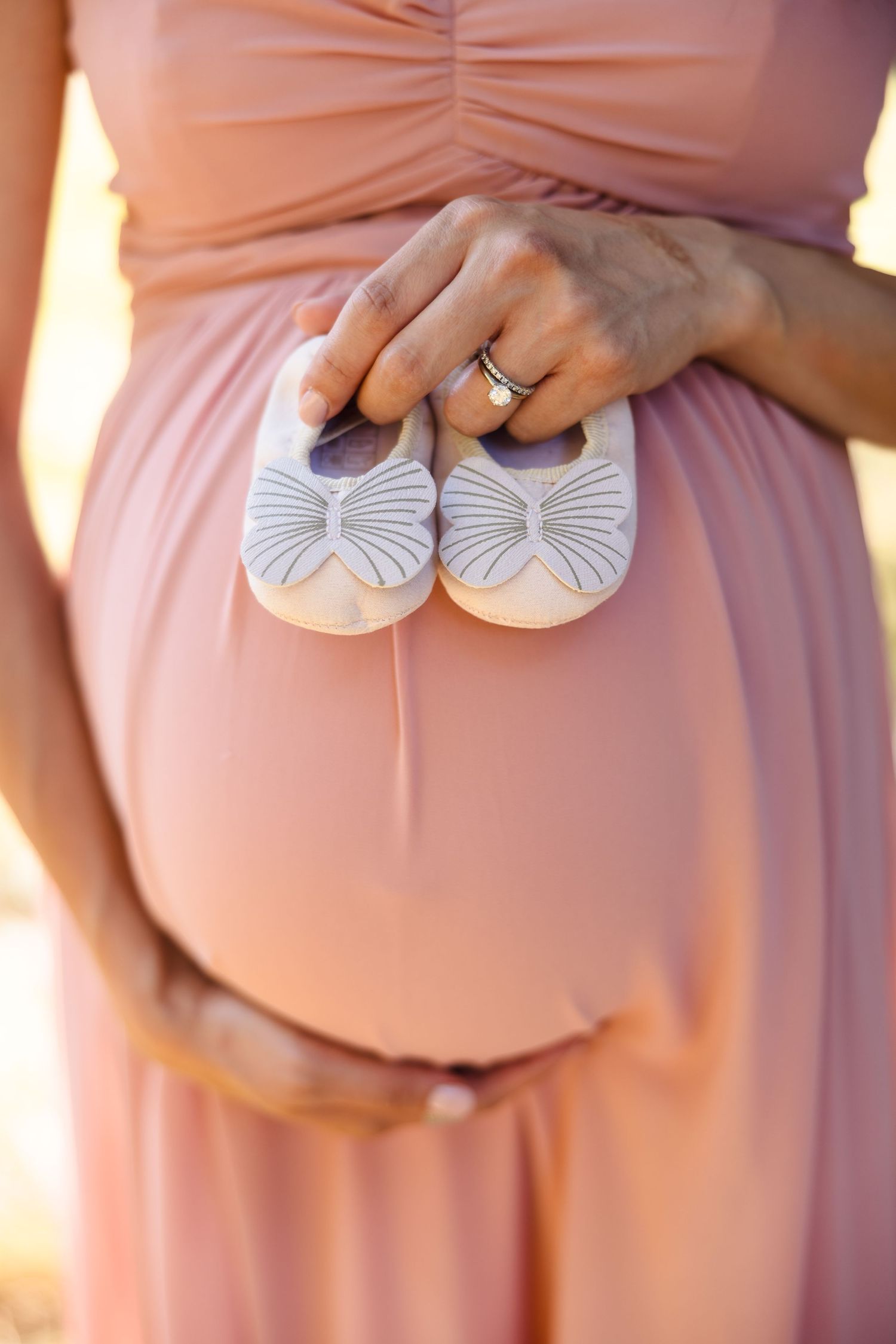 A pregnant woman in a pink dress holds tiny white baby shoes with bow details while cradling her baby bump.