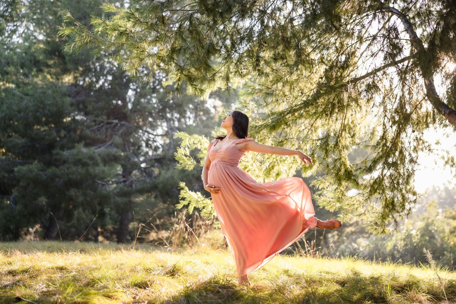 A dancer in a flowing peach dress twirls gracefully in a sunlit forest setting during golden hour.