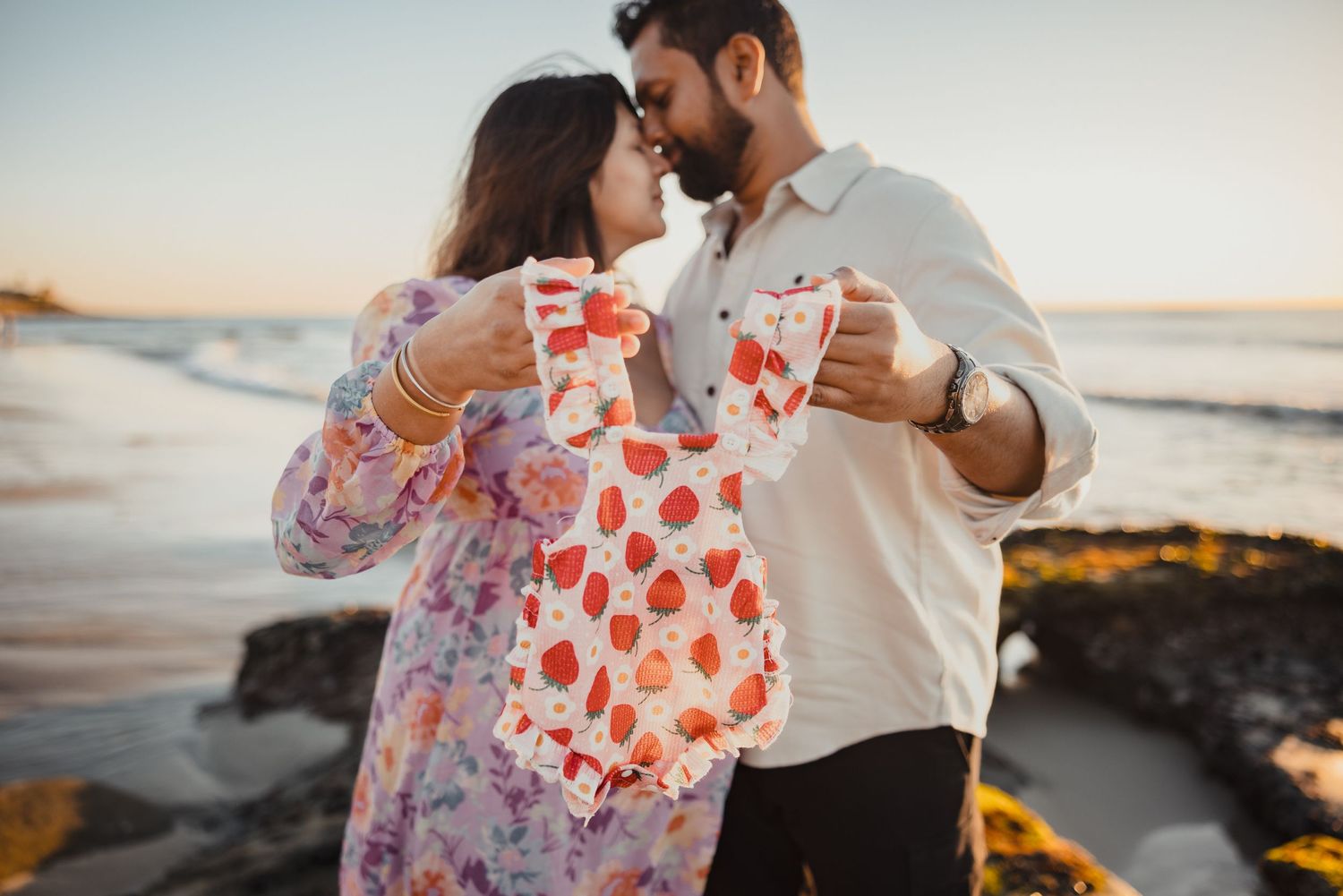 A couple shares drinks on the beach at sunset while wearing floral dress and white shirt outfits.