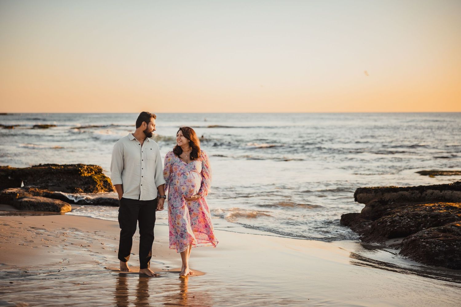 Two people share a romantic moment walking along a rocky beach at sunset as waves crash on the shore.