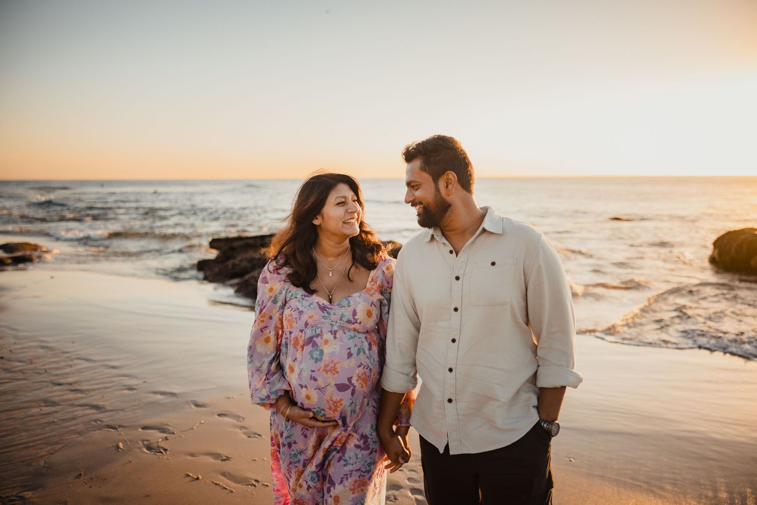 A couple shares a tender moment while walking along a rocky beach at sunset, with golden light illuminating the scene.