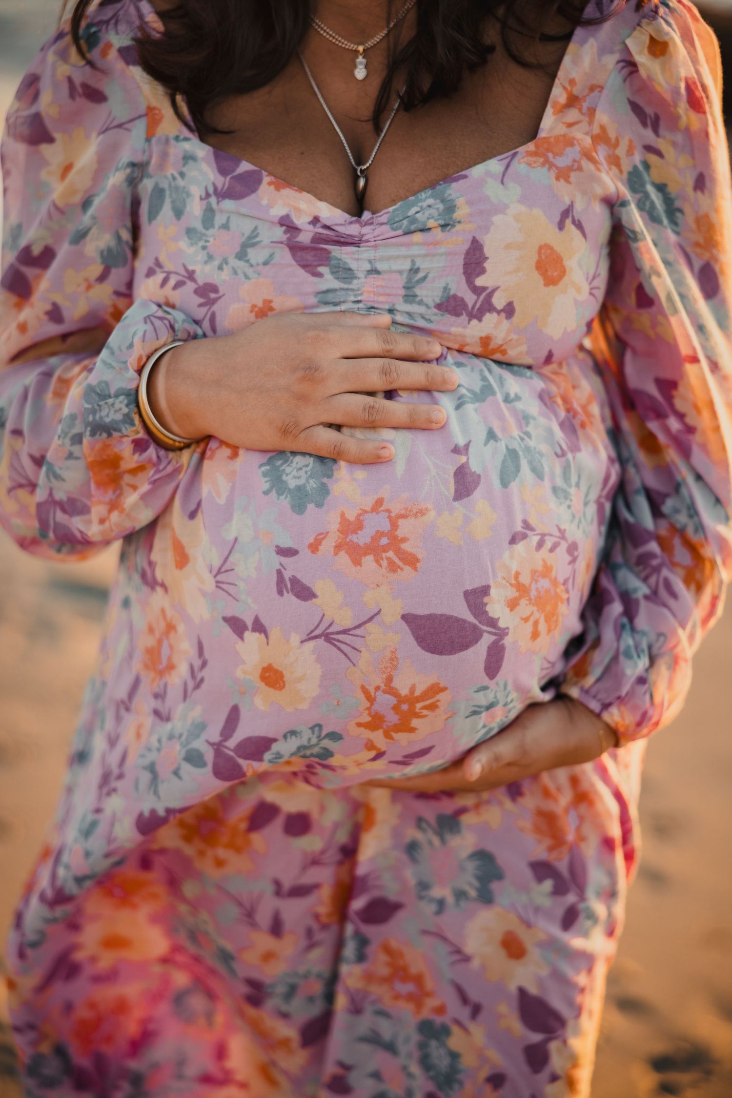 Close up of floral maternity dress with hands gently resting on a pregnant belly at sunset.