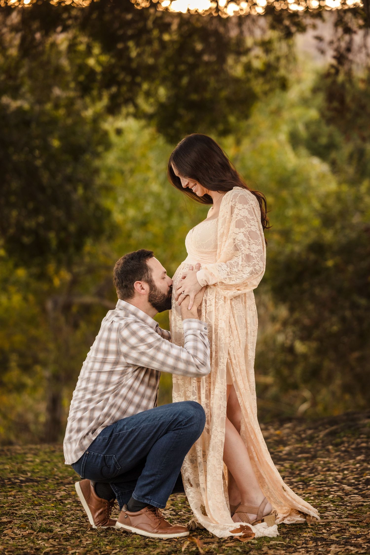 Intimate maternity photoshoot in nature showing couple in tender moment at sunset with lace dress and plaid shirt.