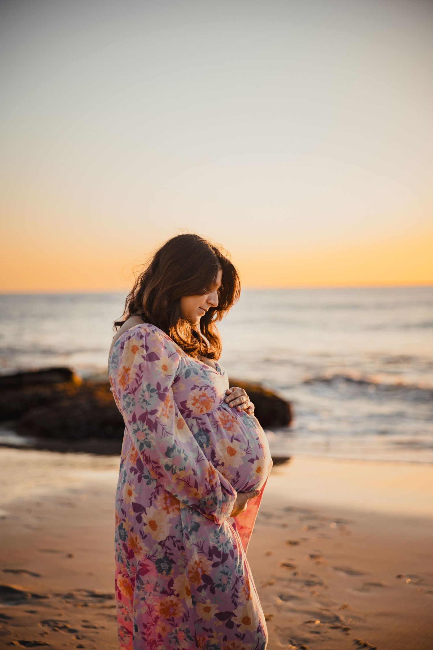 A pregnant woman in a floral dress stands on a beach at sunset, cradling her baby bump.