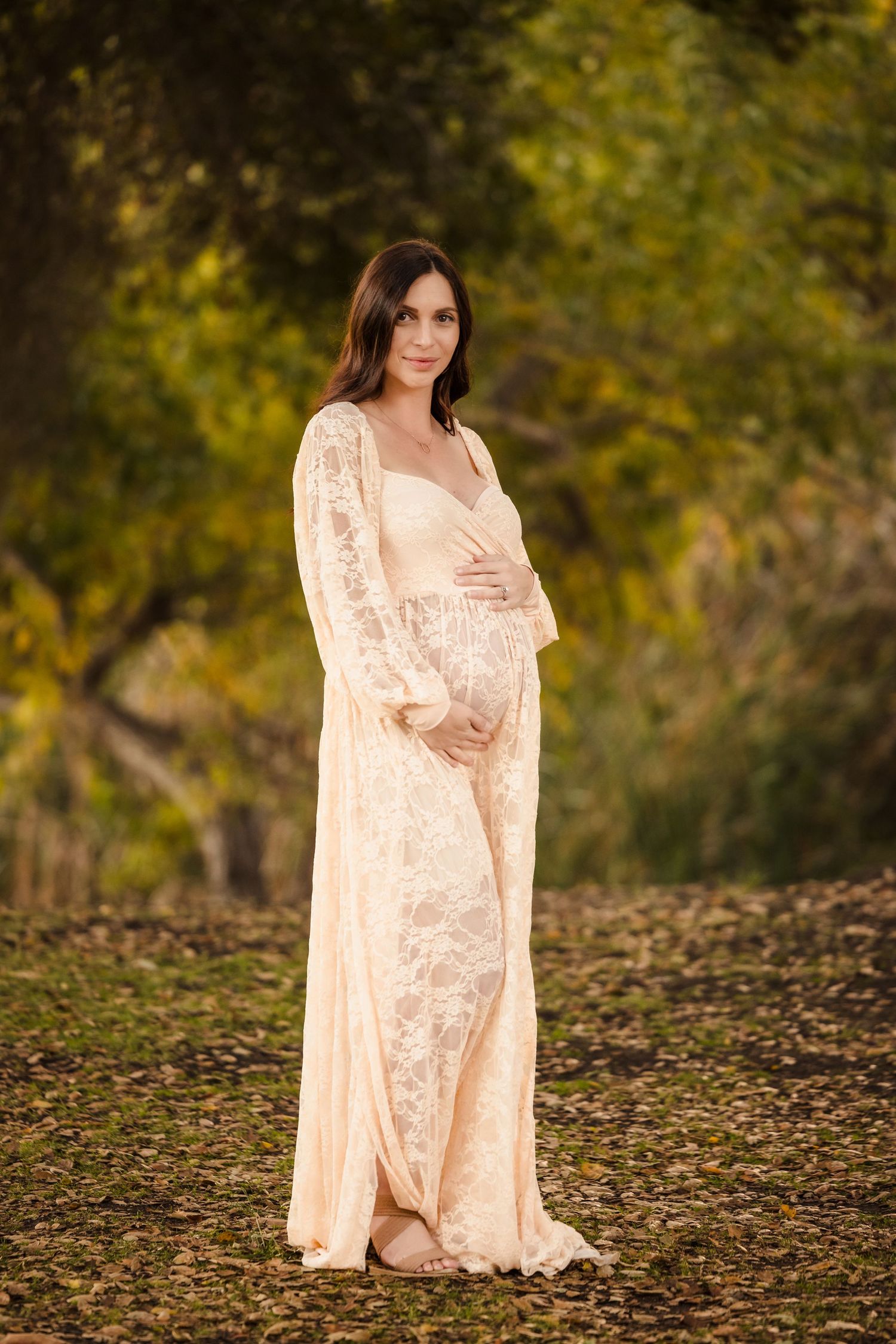 Elegant maternity photo featuring a flowing cream lace dress in an outdoor autumn setting with golden foliage.